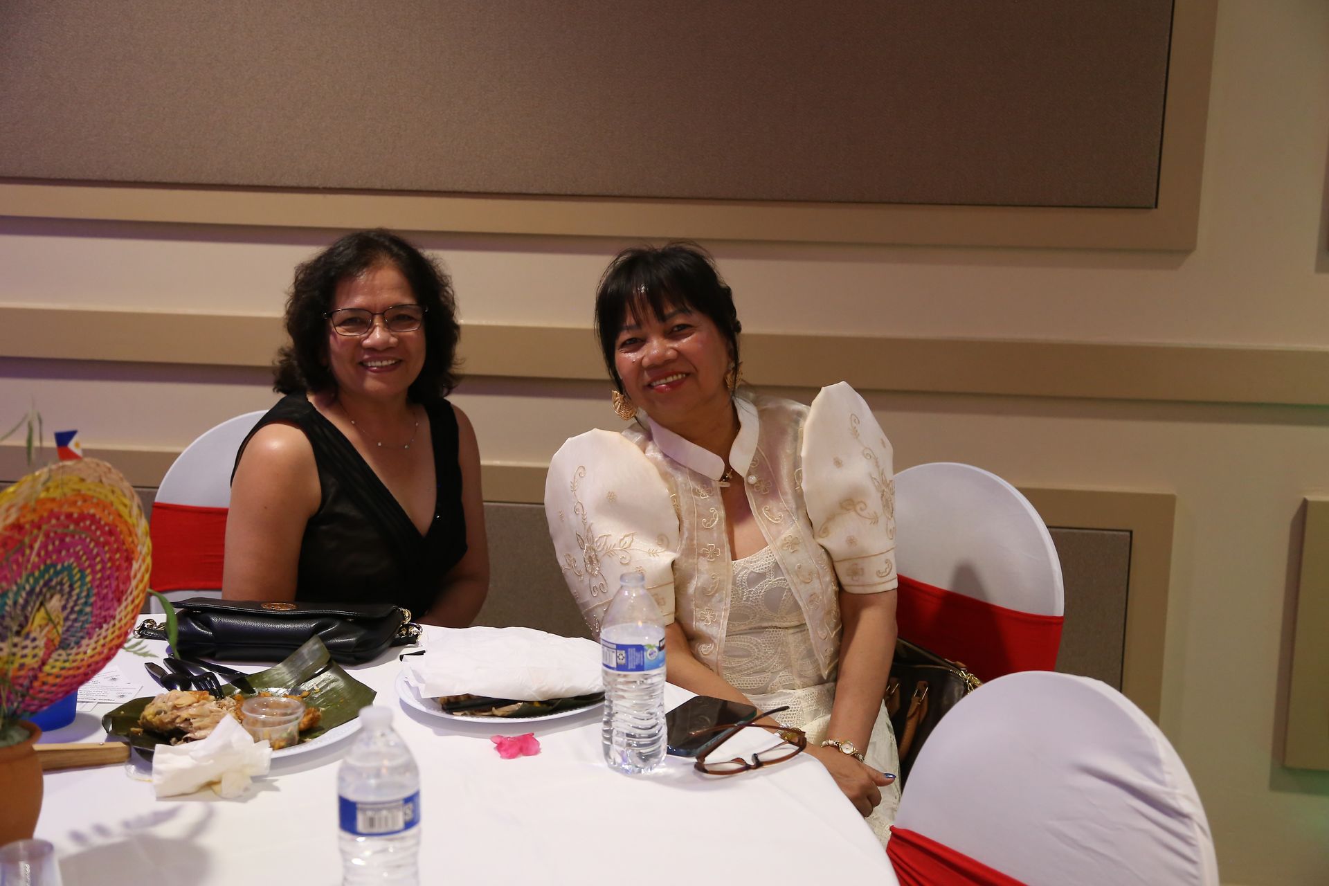 Two people seated at a banquet table, smiling, with plates, drinks, and red chair covers.