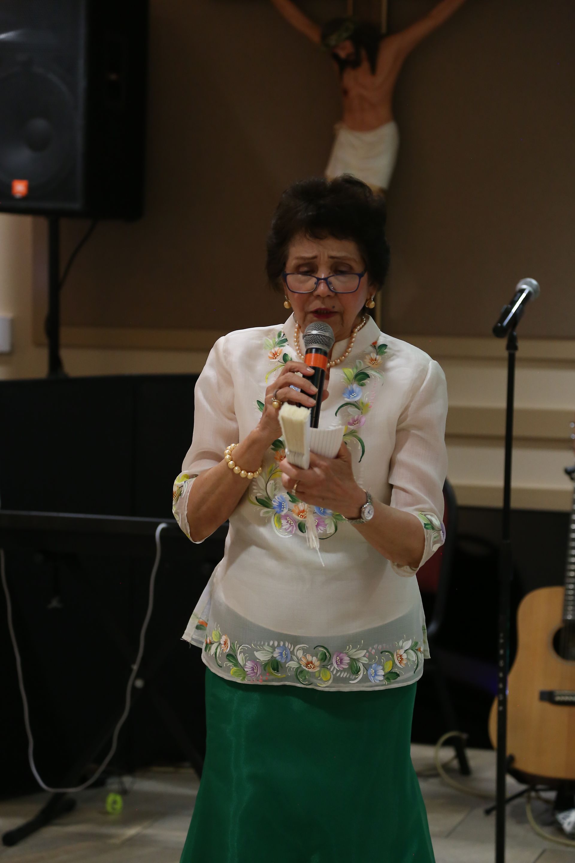 Woman speaking into a microphone on a stage, wearing a white top with floral trim and green skirt.