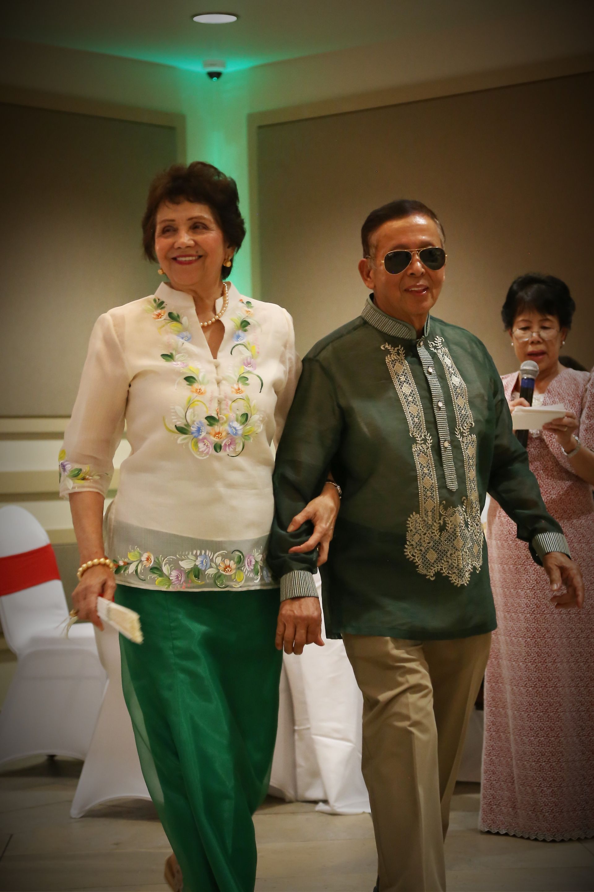 Two people in green traditional outfits walking indoors at a formal event
