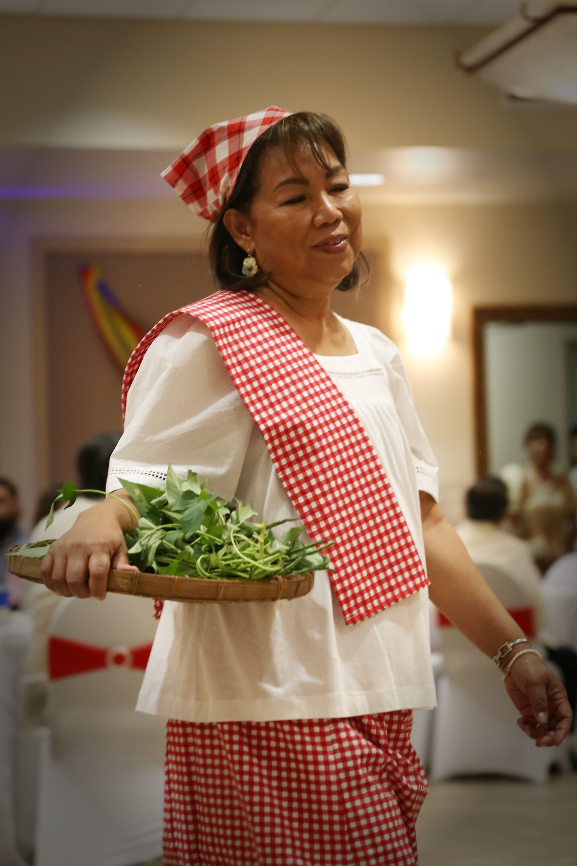 Woman in red-checkered outfit carrying a basket of greens at an indoor event