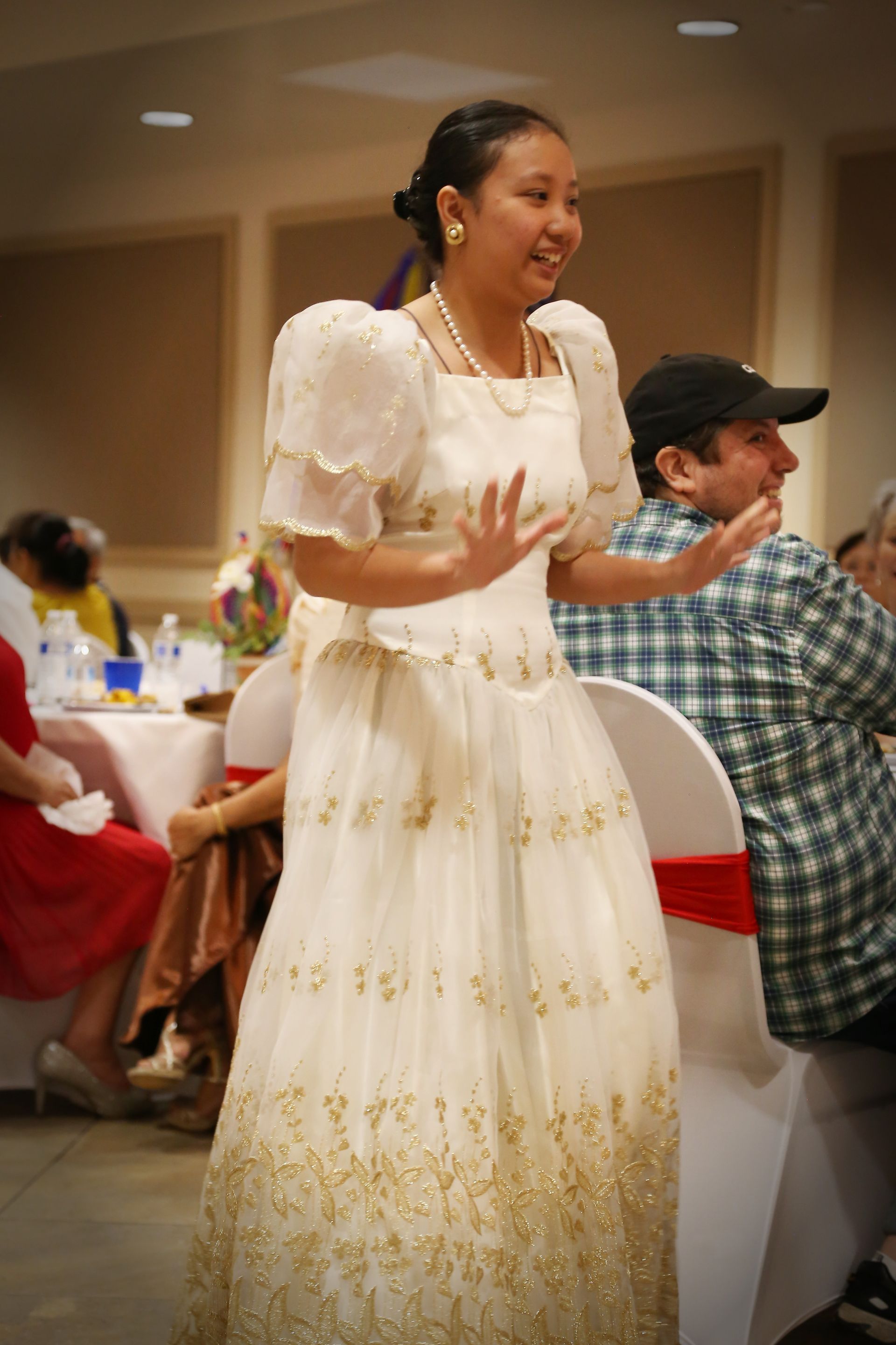 Woman in a white dress speaking animatedly at a banquet table in a warmly lit hall