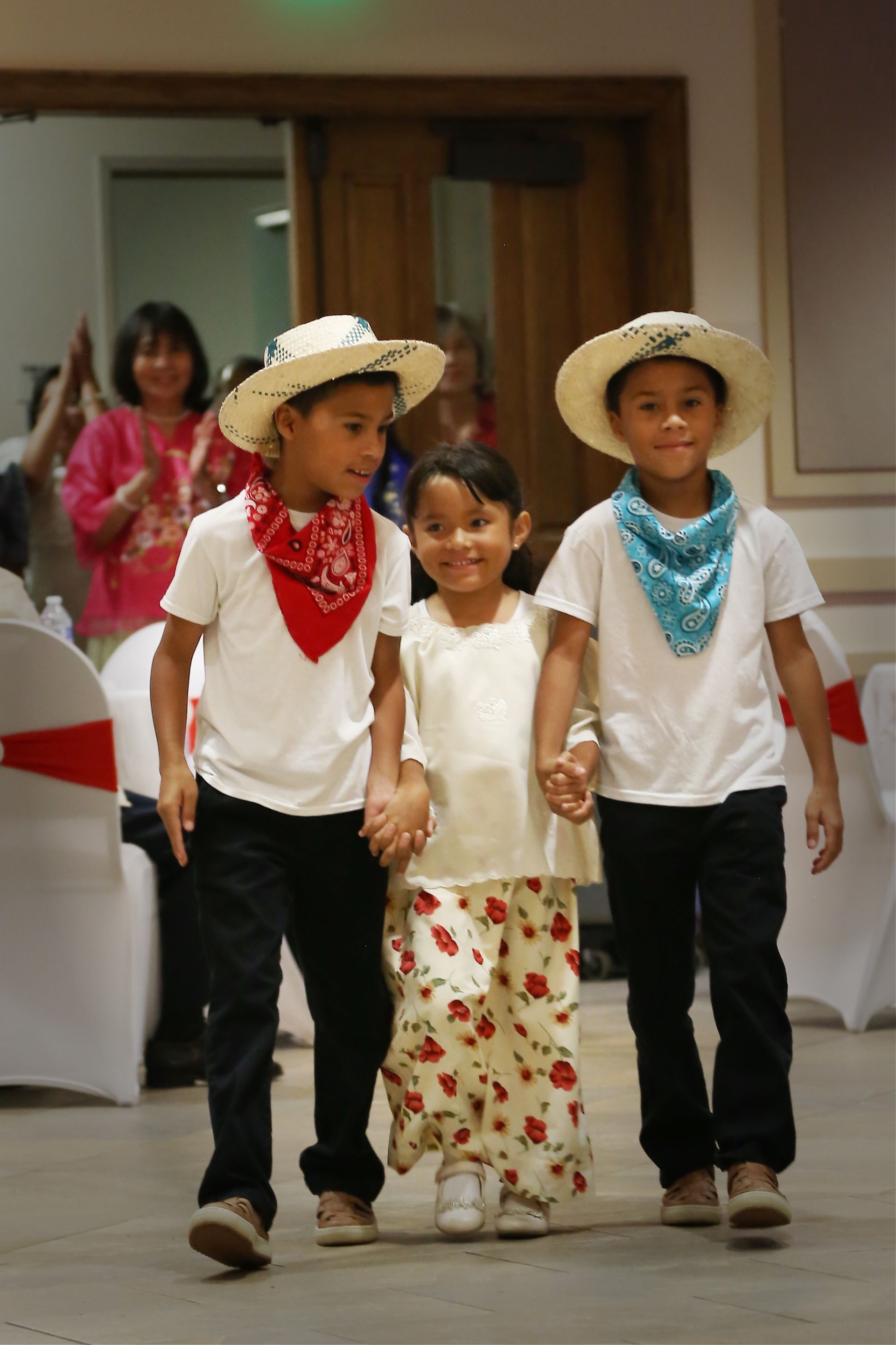 Three children in straw hats and bandanas walking indoors, two boys holding hands with a girl between them
