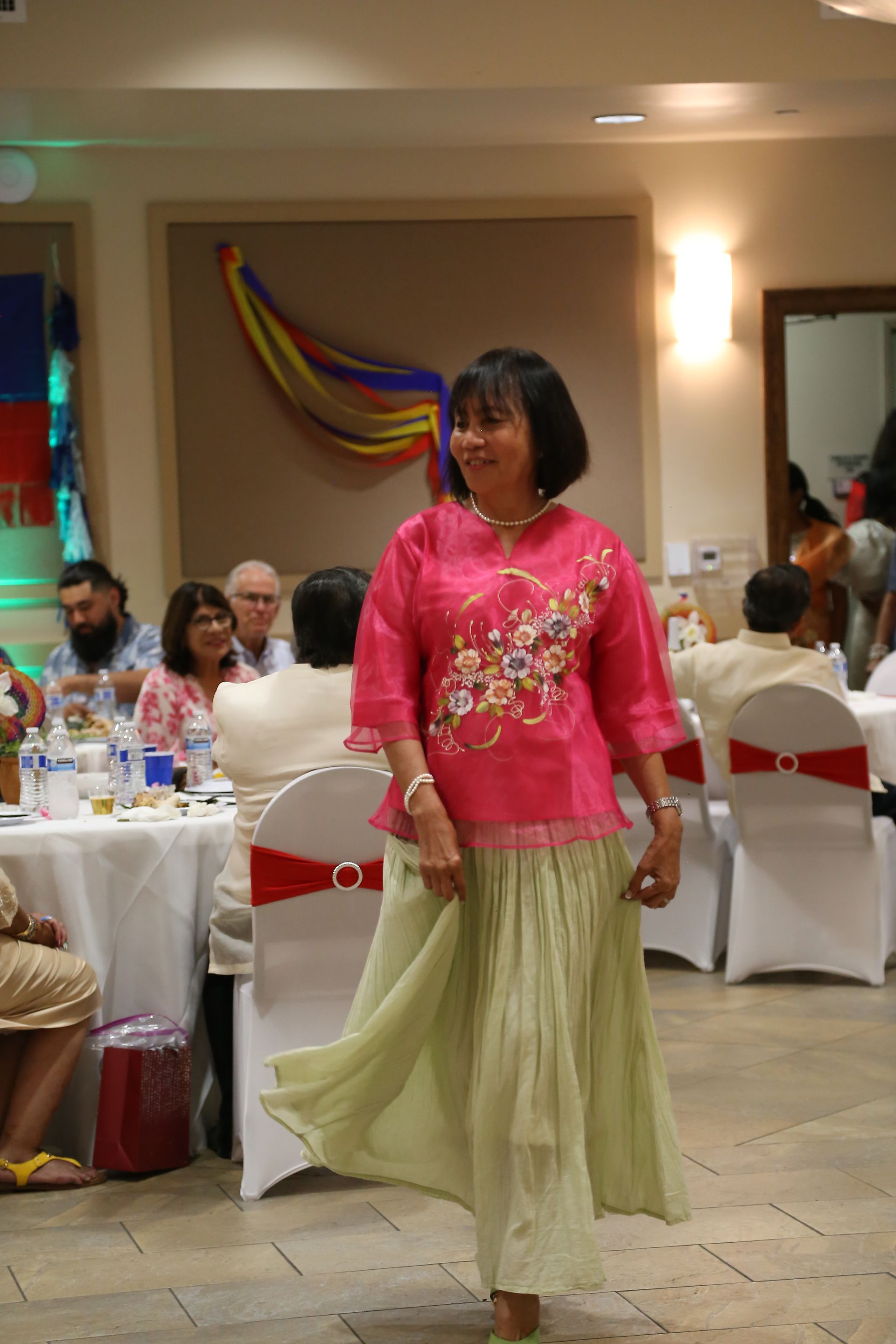 Woman in pink blouse and green skirt walking at a decorated indoor event banquet.