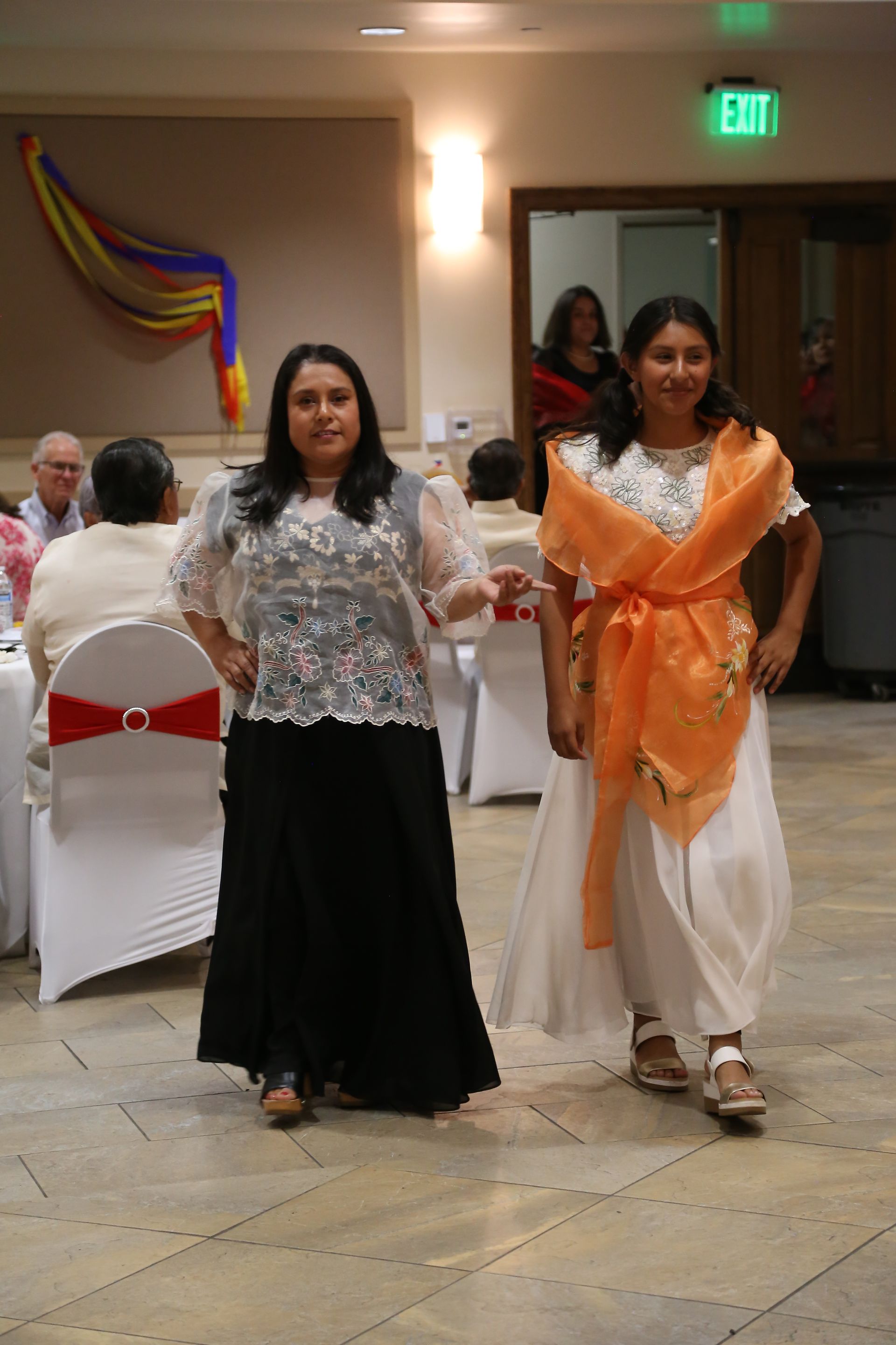 Two women walk through a banquet hall in dressy outfits, with seated guests and a green exit sign behind them.