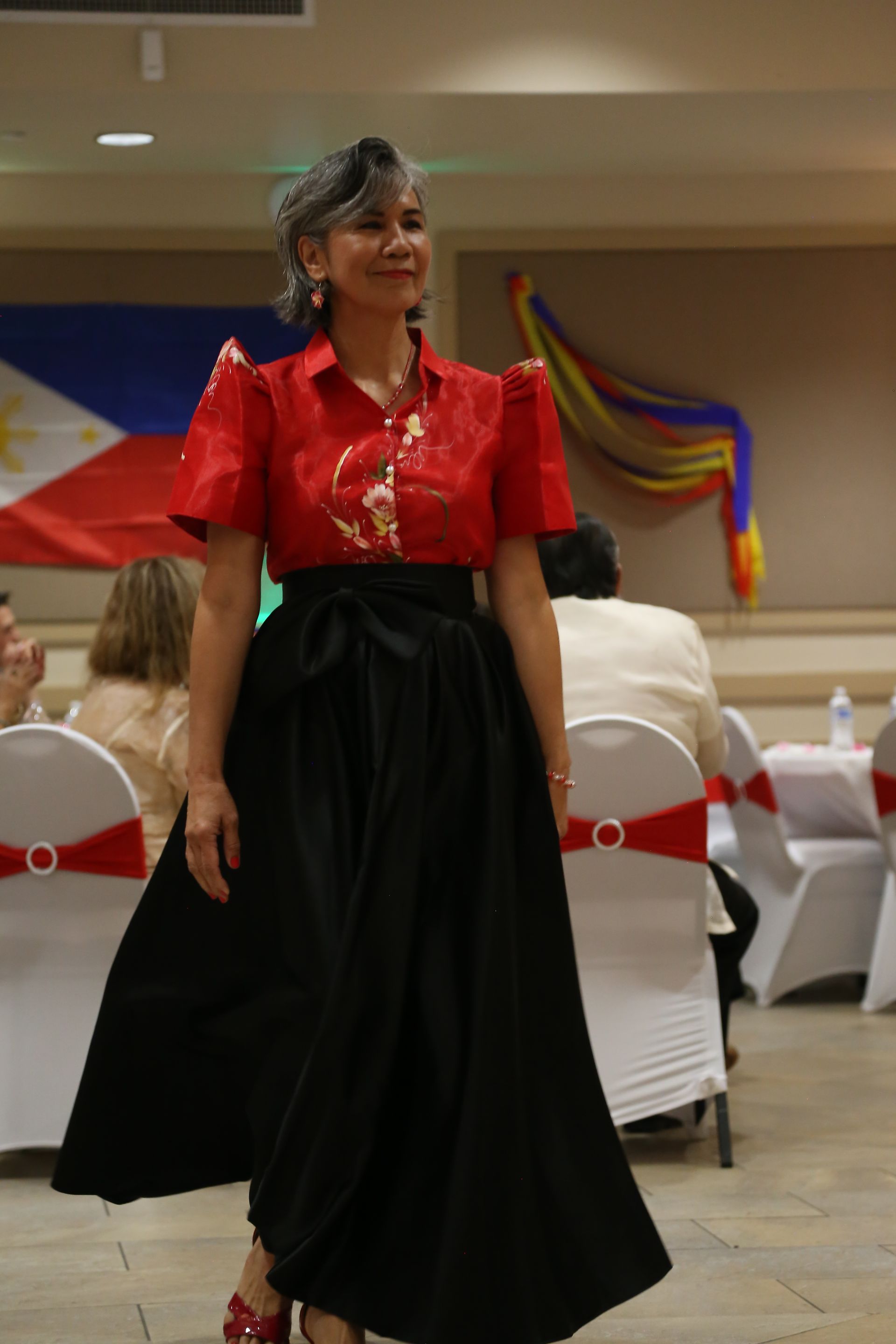 Woman in red blouse and black skirt walking through a decorated banquet hall with white chair covers and flags.