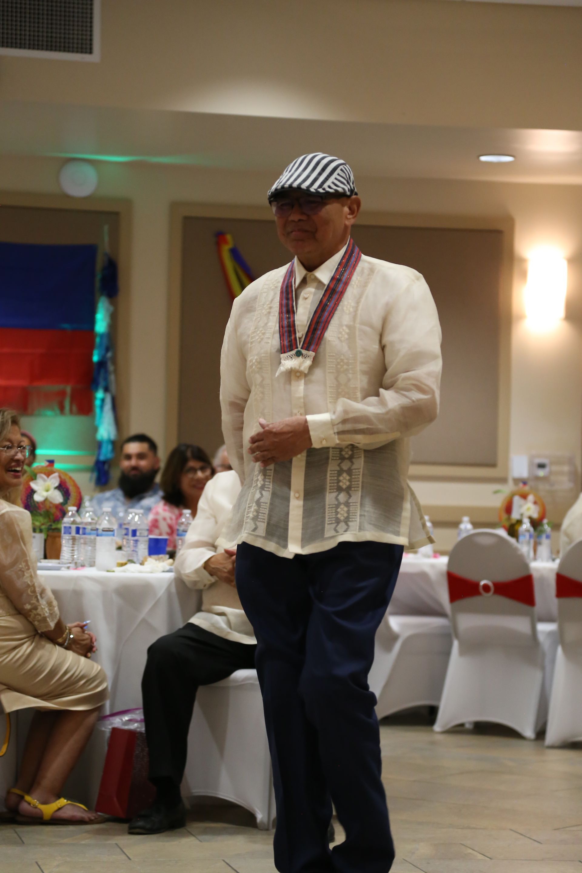 Man in a cream shirt and striped cap standing at a banquet table in a decorated hall