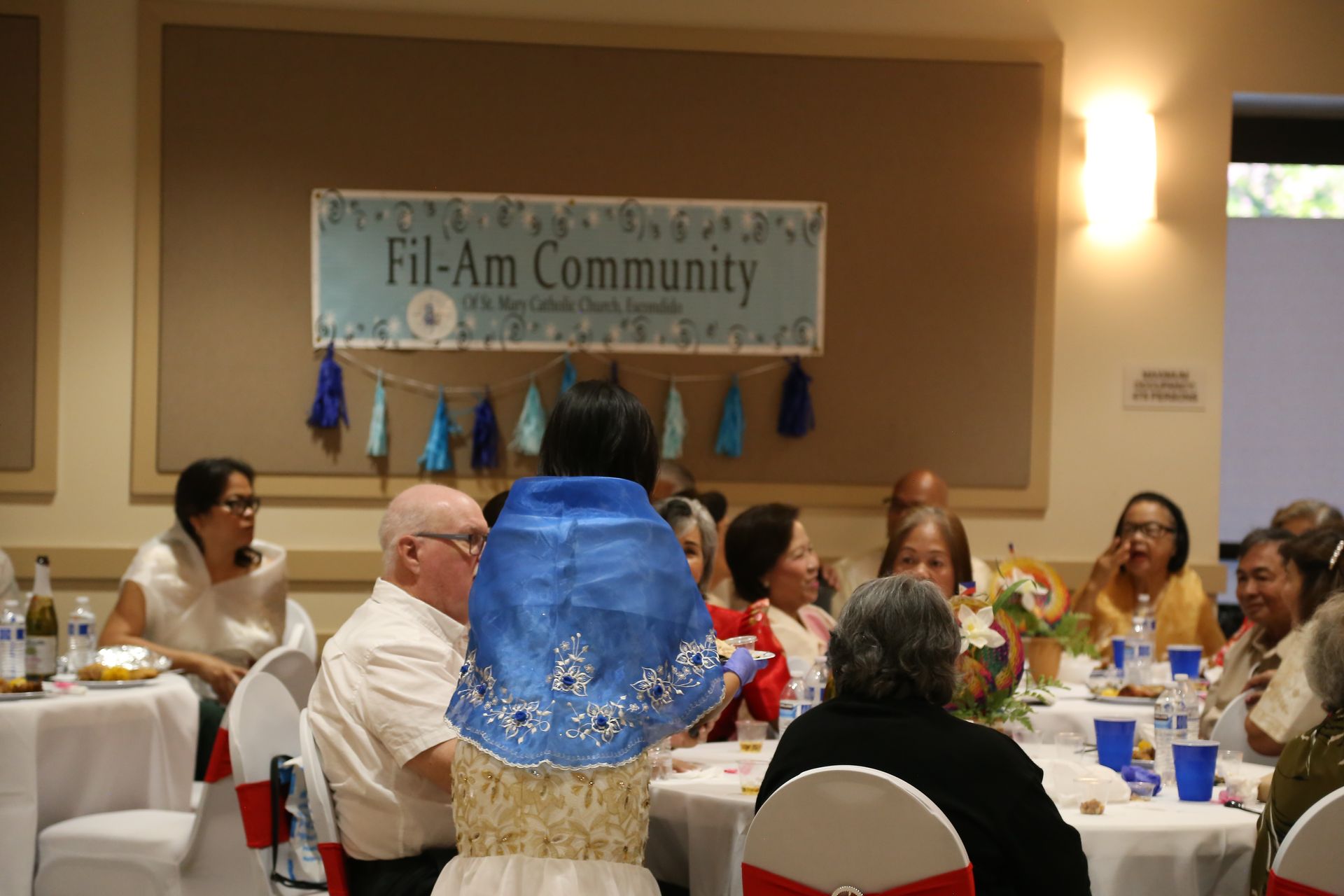 Community banquet with guests seated at tables, a person in a blue dress in the foreground, and a “Fil-Am Community” banner.
