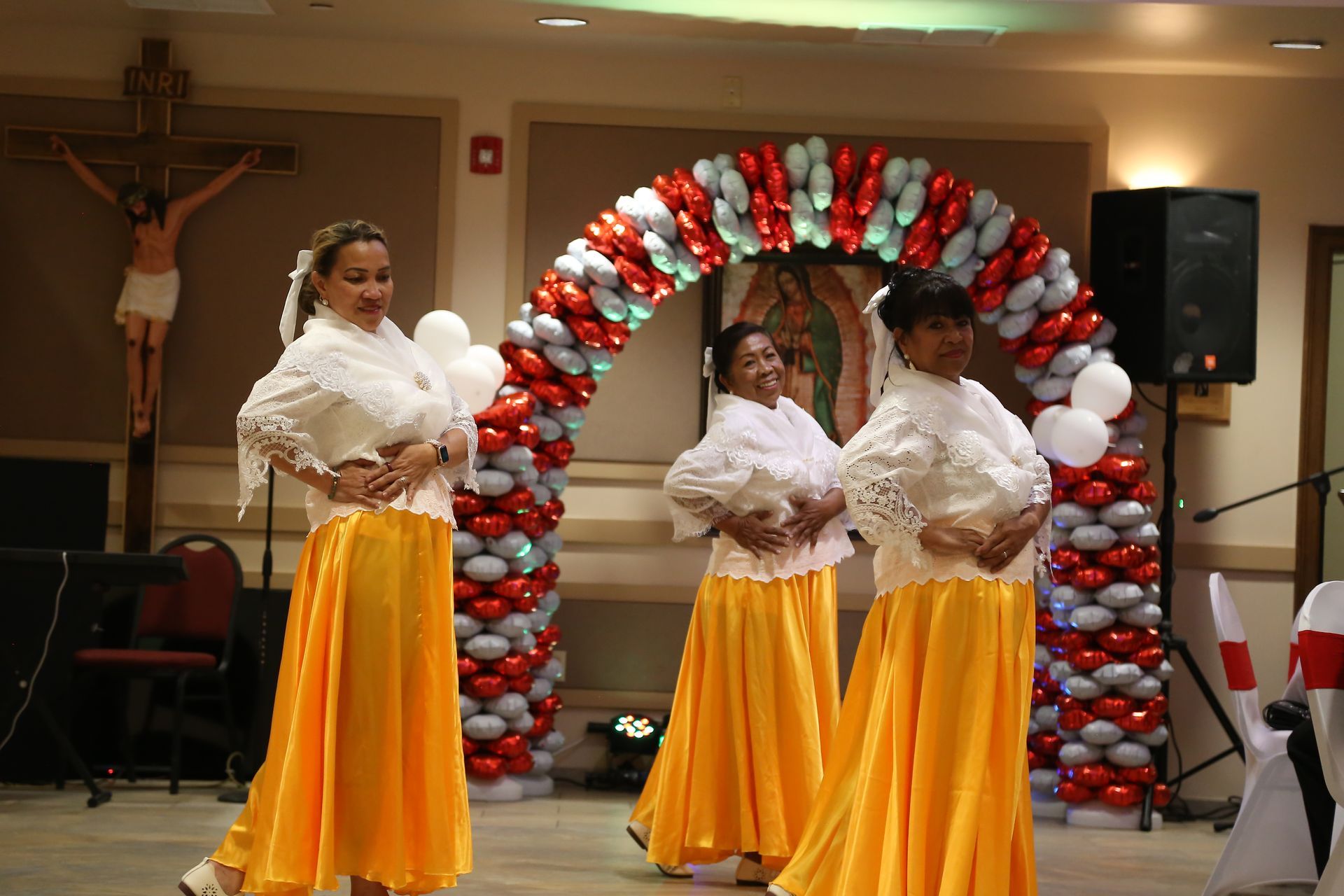 Three performers in yellow skirts and white ruffled tops dance beneath a red-and-white balloon arch.