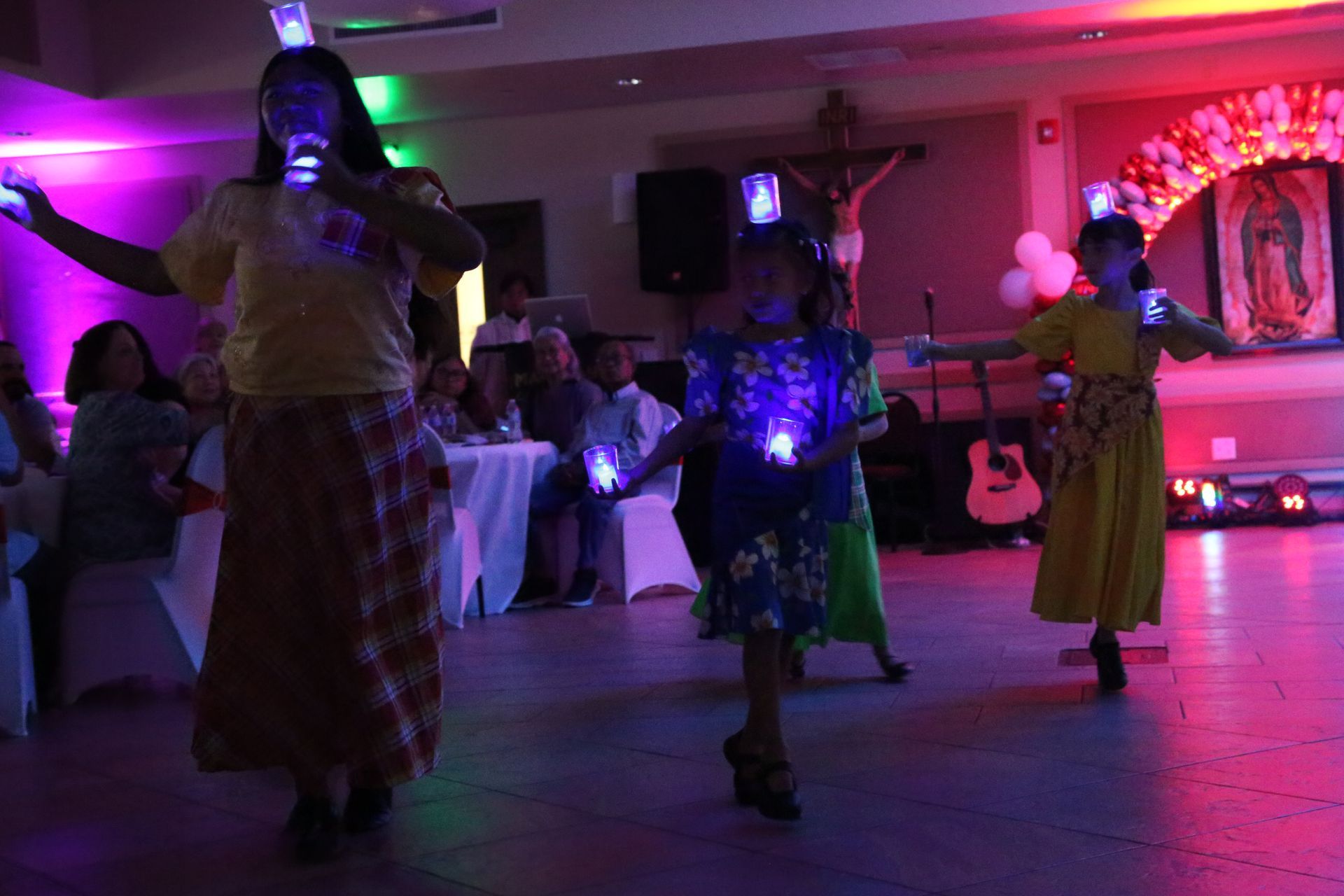 Three costumed dancers perform under purple lights in a banquet hall with seated guests.
