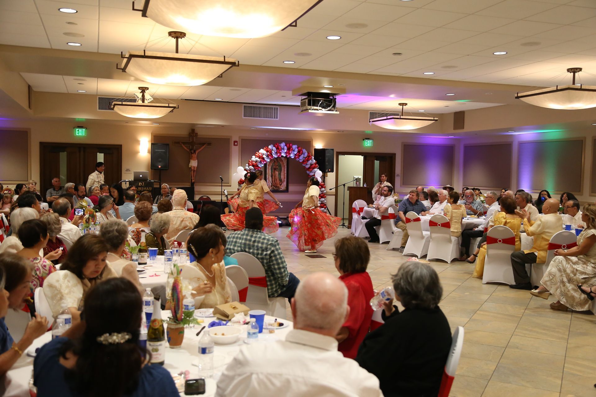Banquet hall reception with guests seated at round tables and a couple under a flower arch dancing