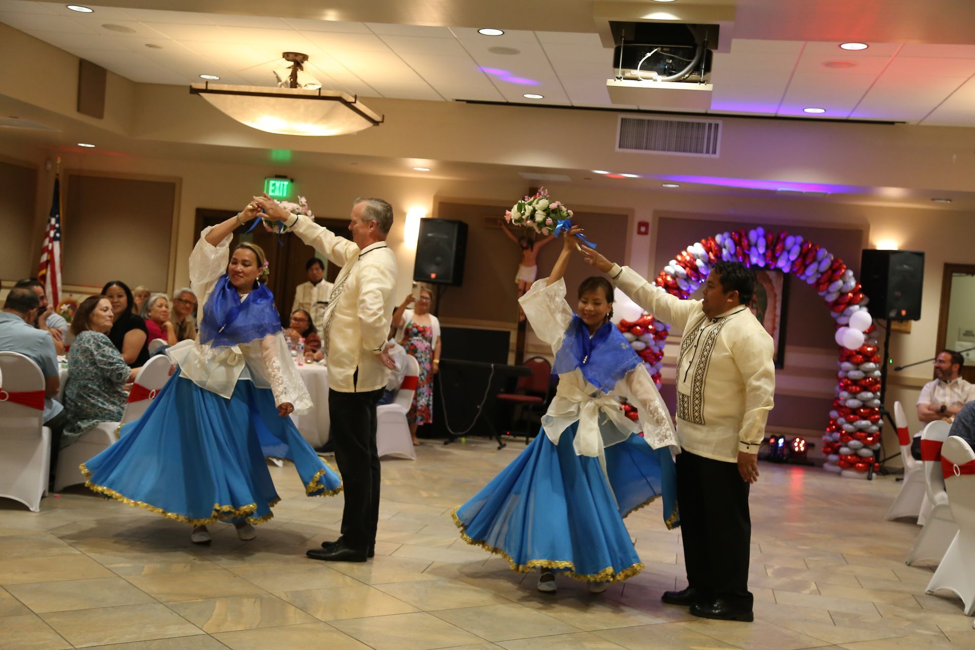 Four dancers in blue skirts perform in a decorated banquet hall, with two men and two women in beige shirts.