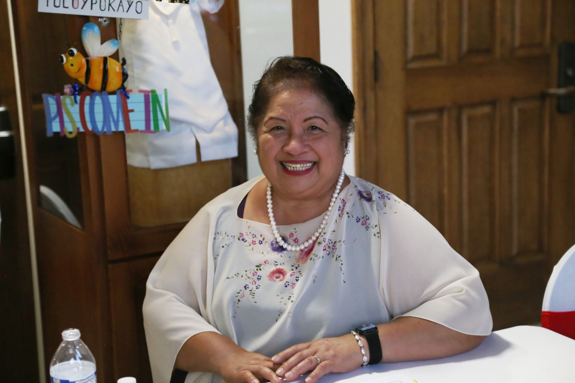 Smiling woman seated at a table in a wooden room, wearing a white blouse and necklace
