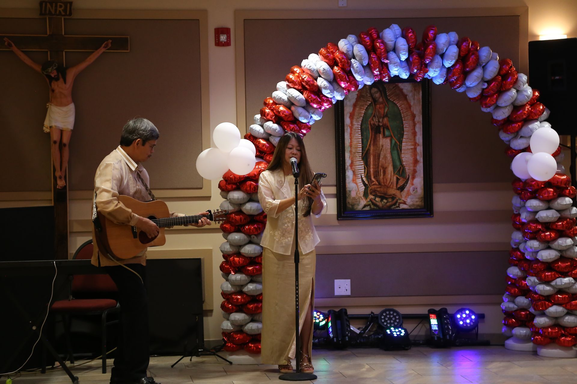 Singer at a microphone beside a guitarist on a stage under a red, white, and blue balloon arch