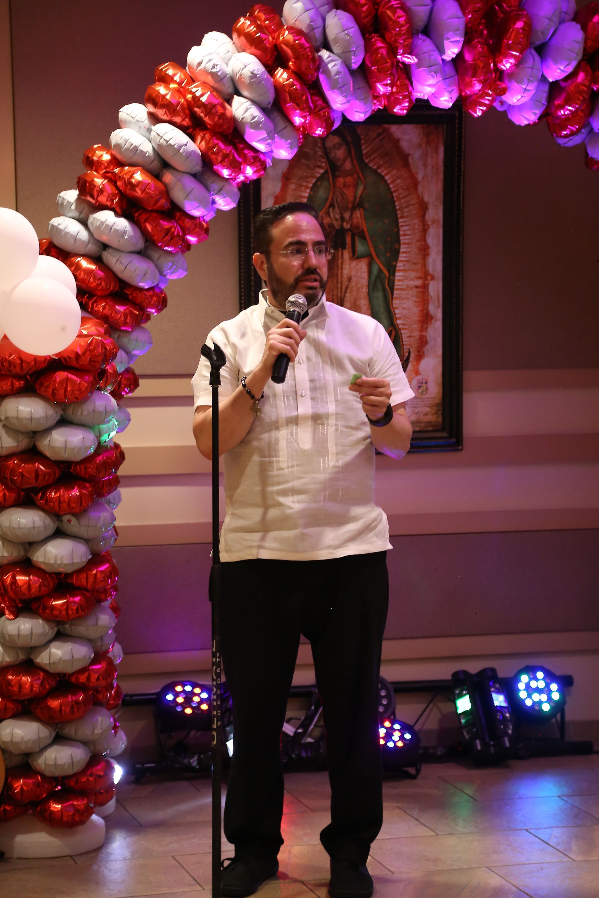 Man speaking into a microphone under a red-and-white balloon arch at an indoor event