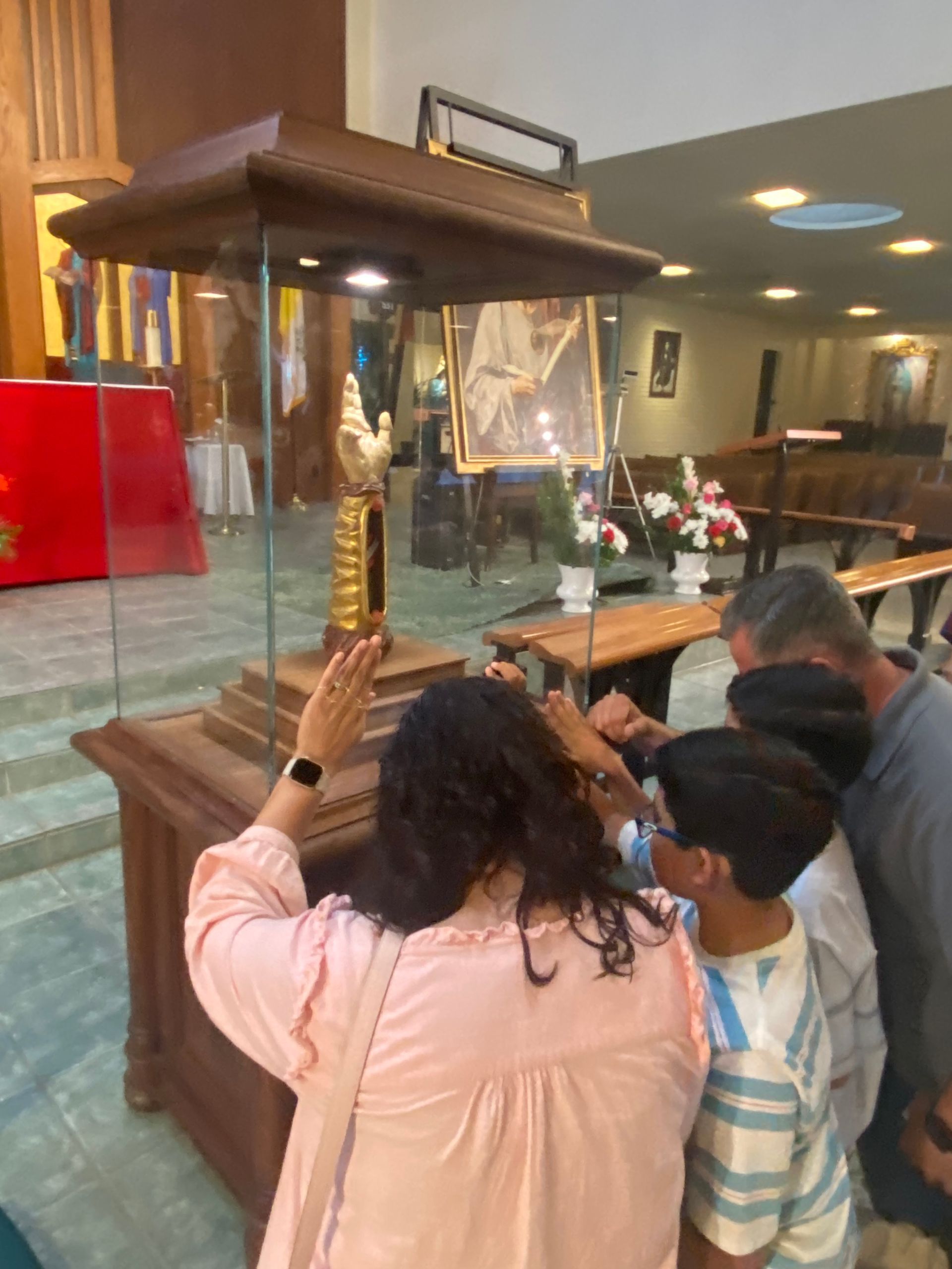 People praying at a glass-encased reliquary in a church, with candles and flowers nearby
