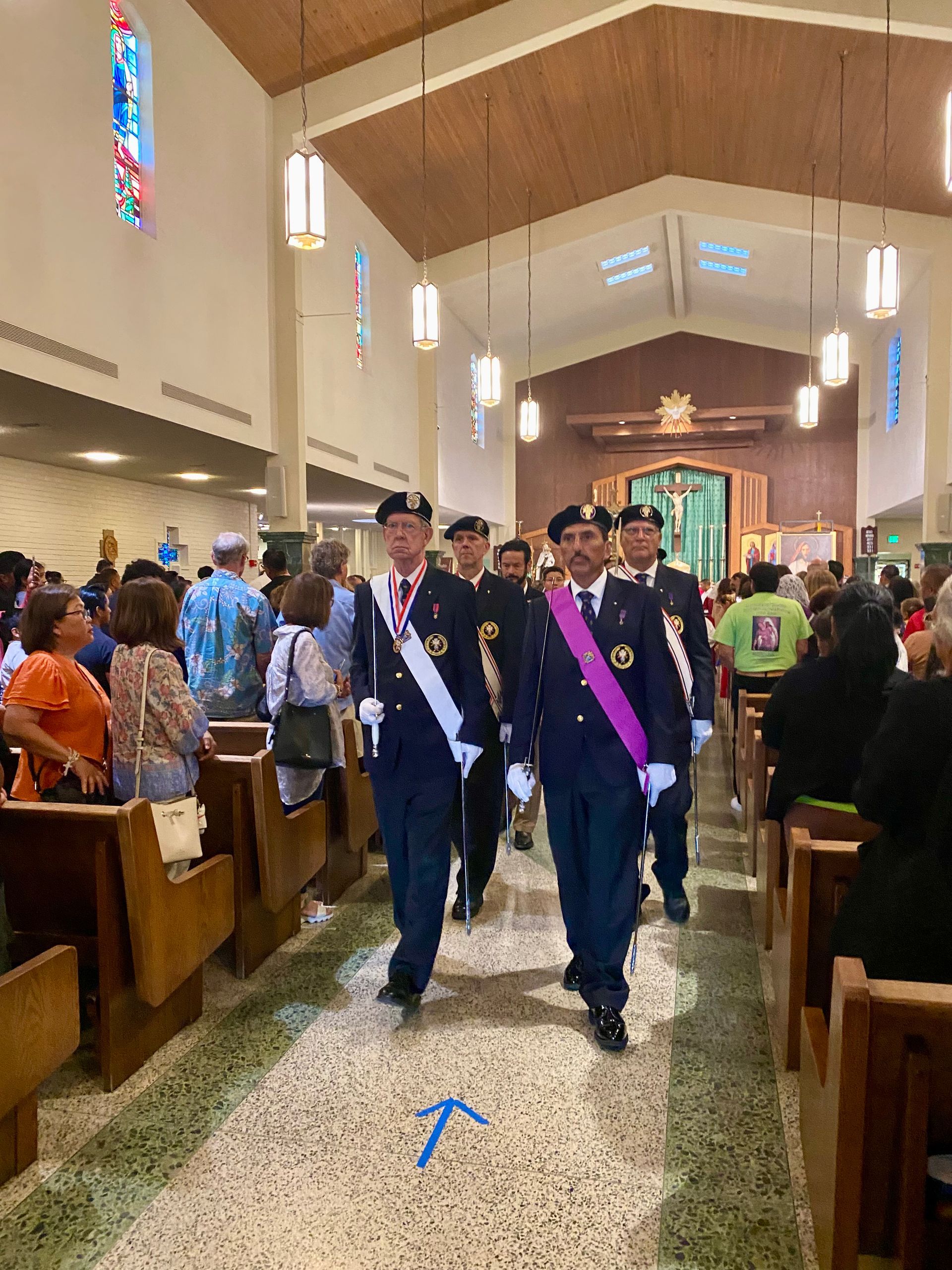 Two men in dark suits and sashes walk down a church aisle during a ceremony