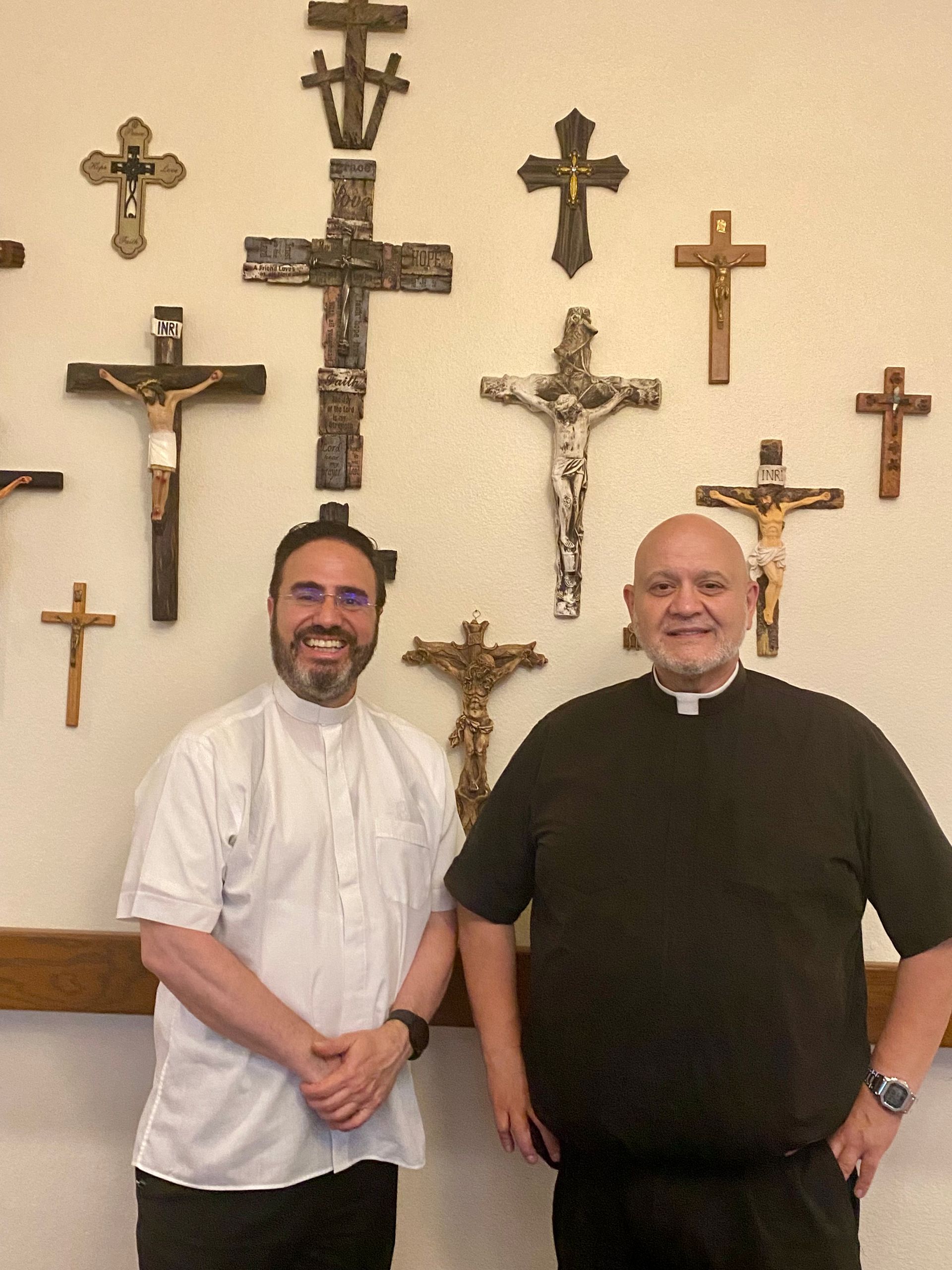 Two men stand smiling in front of a wall covered with crosses.