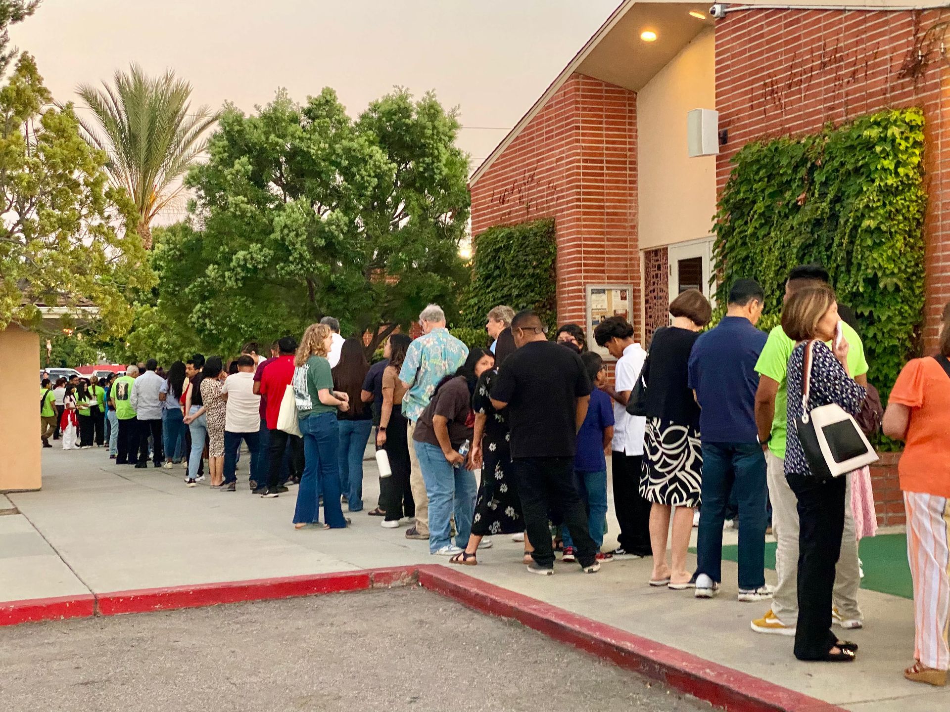People lined up outside a brick building along a sidewalk near a red curb at dusk.