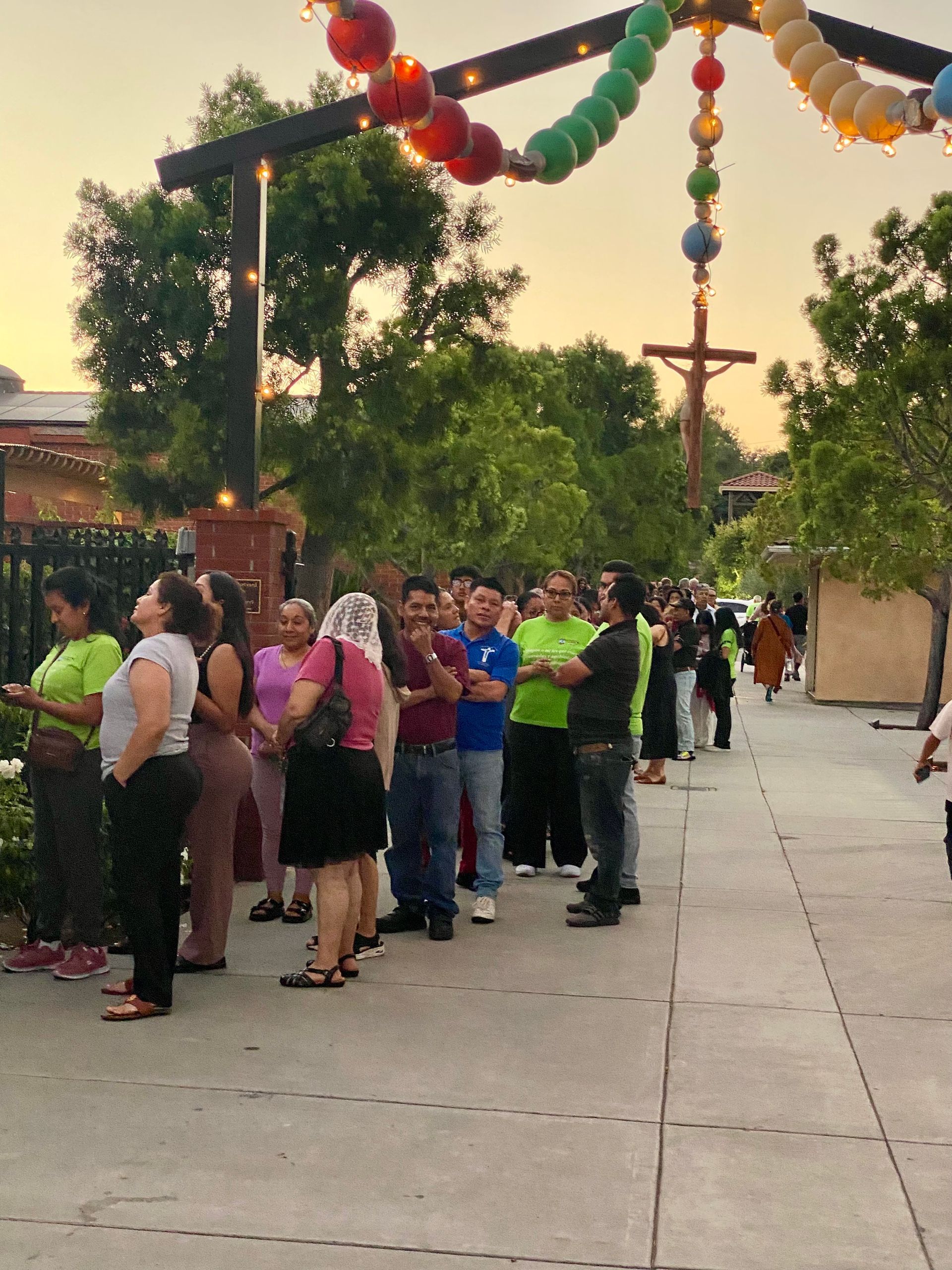 People line up outdoors beneath colorful hanging decorations near a courtyard walkway.