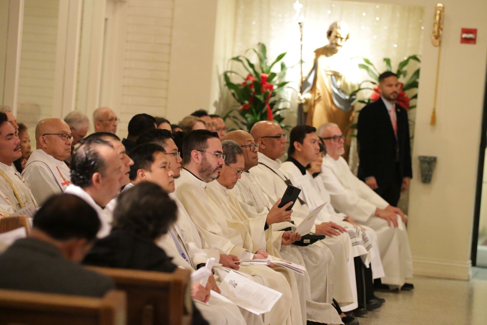 People seated in rows at a formal indoor ceremony, with white robes and floral arrangements nearby