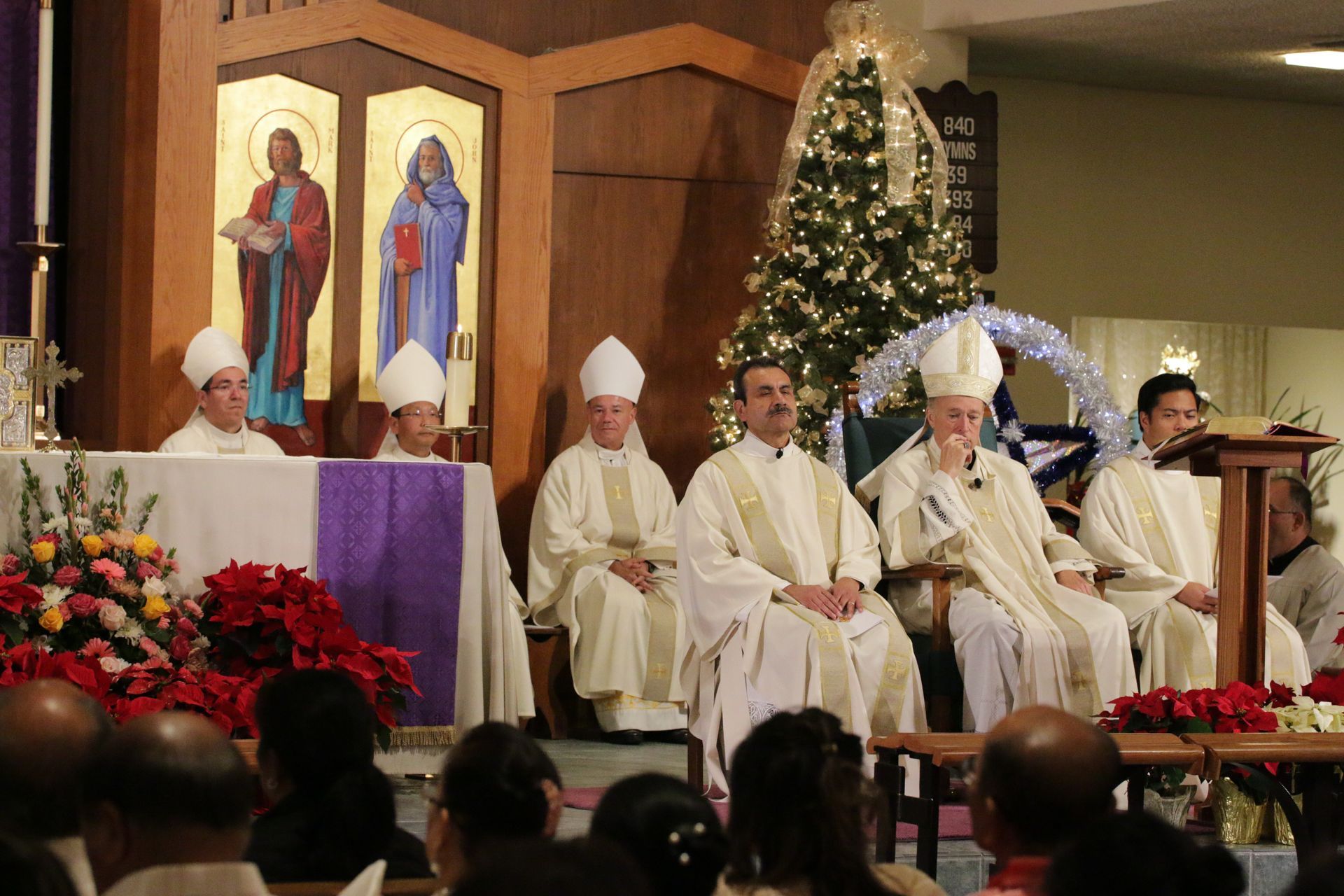 Clergy in white robes seated on a stage beside a lit Christmas tree and religious icons during a церemony