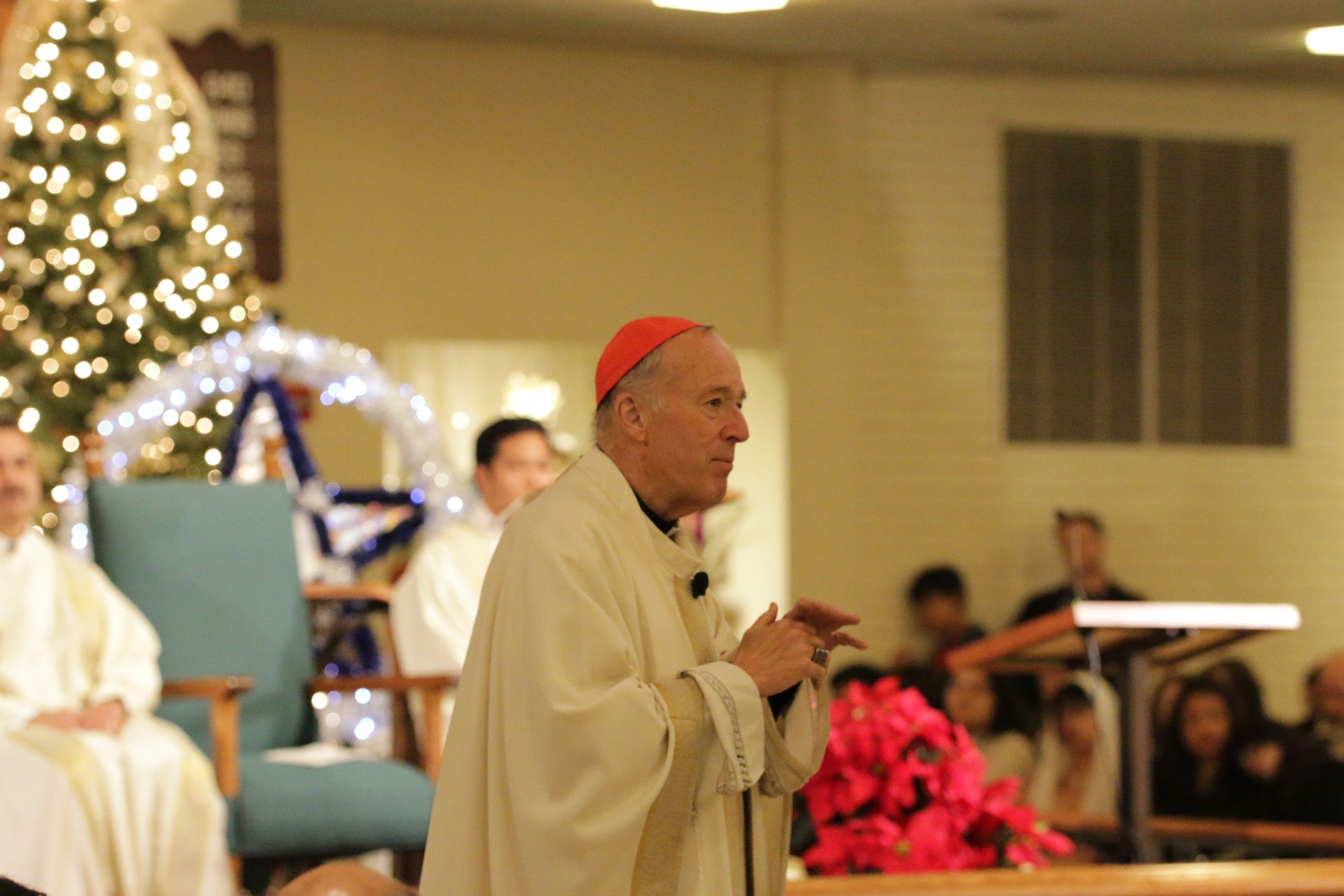 Clergyman in white vestments and red cap stands in a church near a lit Christmas tree, holding a microphone.