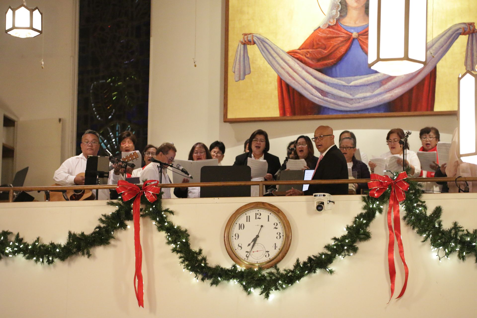 Choir seated on a decorated church balcony, with greenery, red ribbons, and a clock on the wall