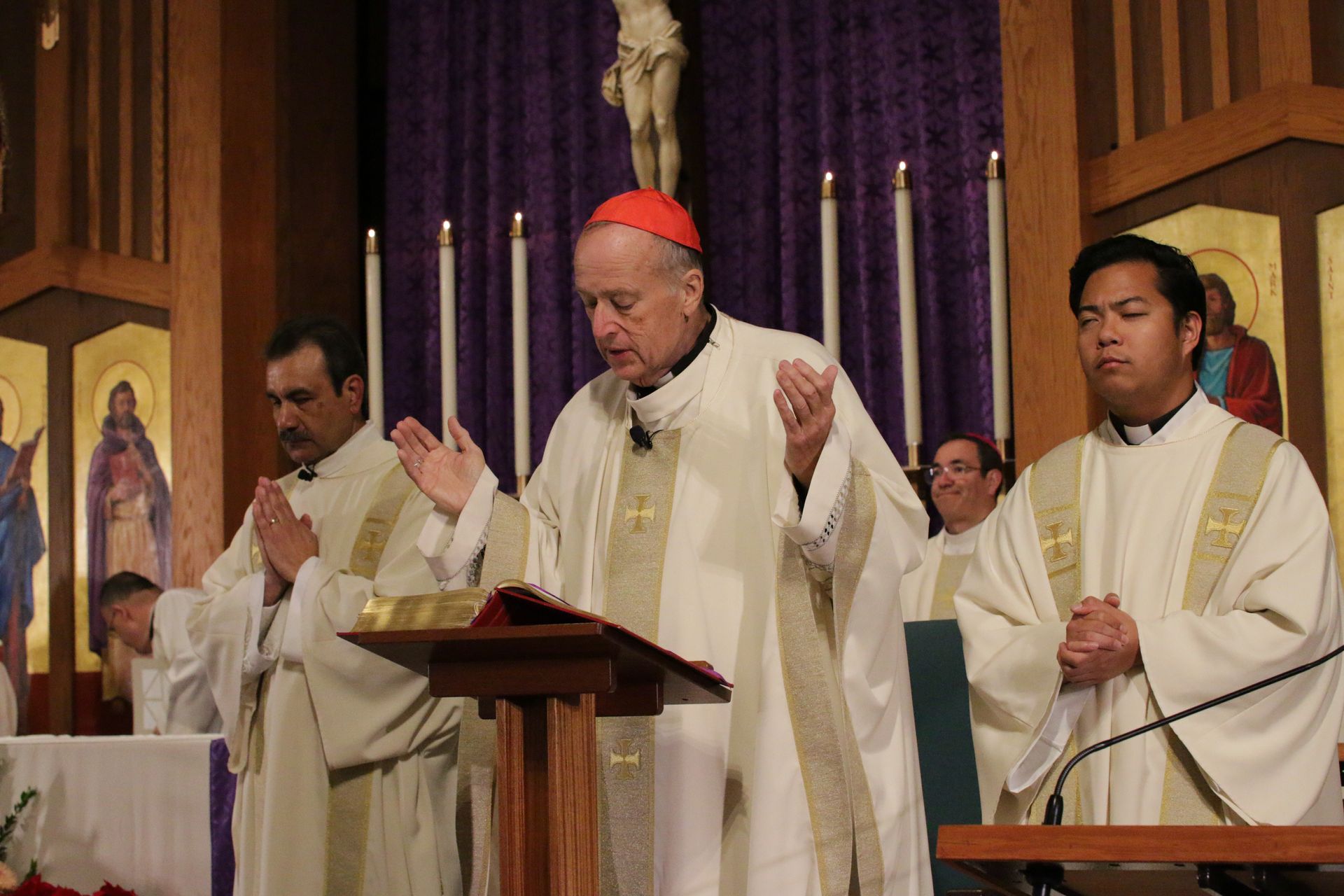 Clergy in white vestments pray at a church altar, with a bishop in red skullcap centered before purple drapery.