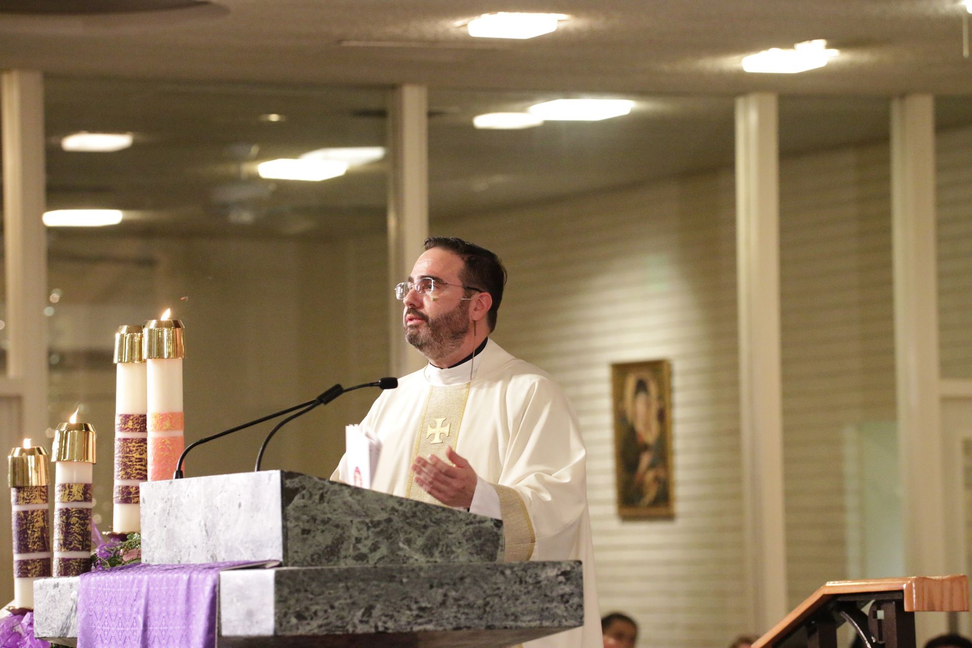 Man speaking at a church lectern during a service, with candles and a crucifix nearby.