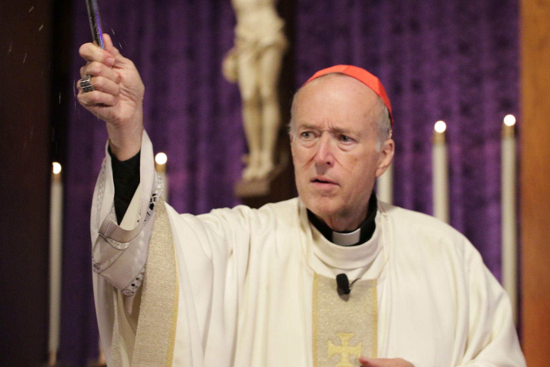 Clergyman in white robes with a red skullcap raising a lit candle in a church interior.