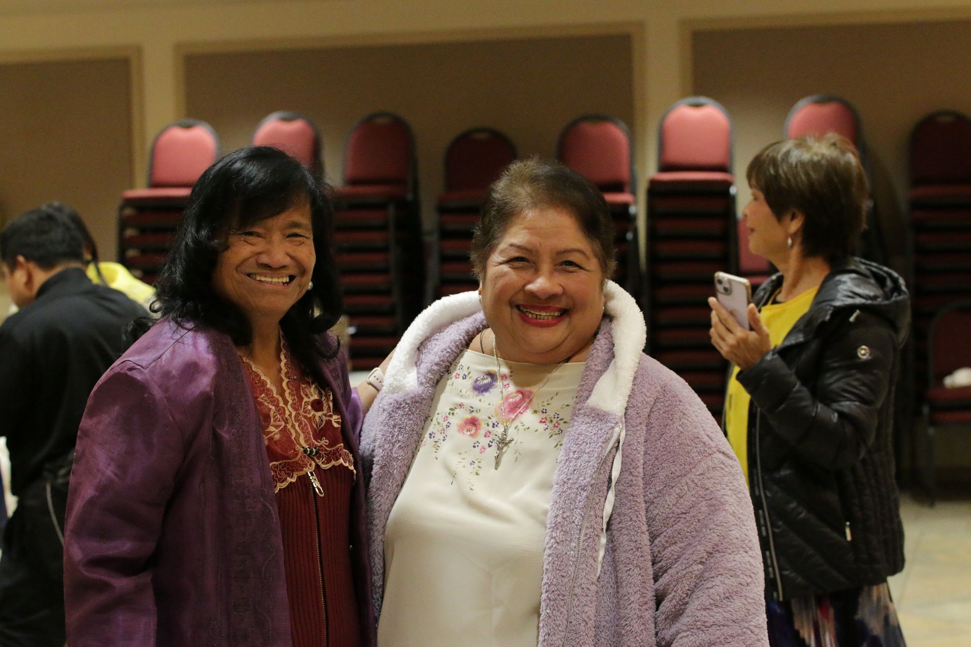 Three women smiling in a room with stacked chairs, one holding a drink.