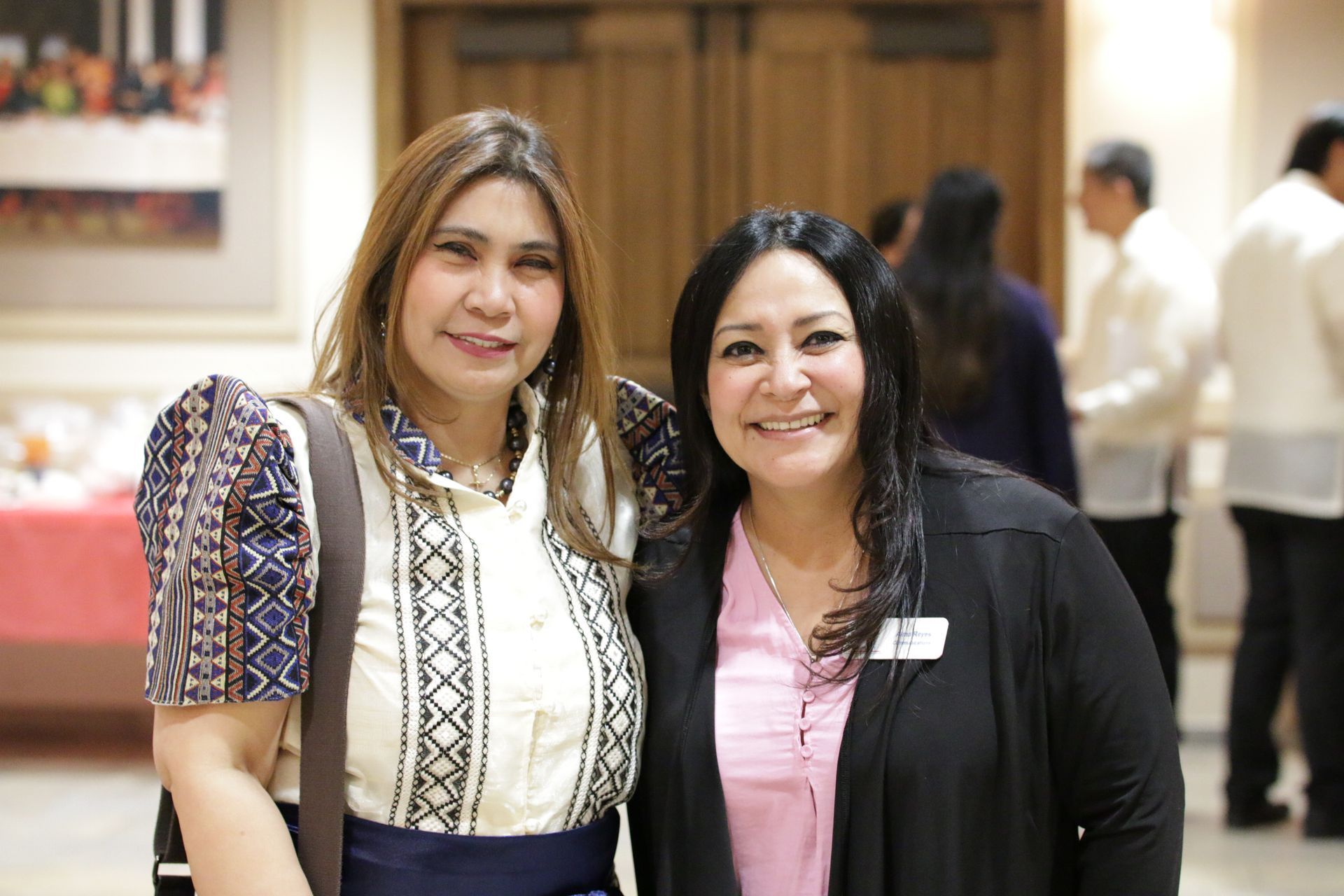 Two women smiling indoors at a social event, standing side by side.
