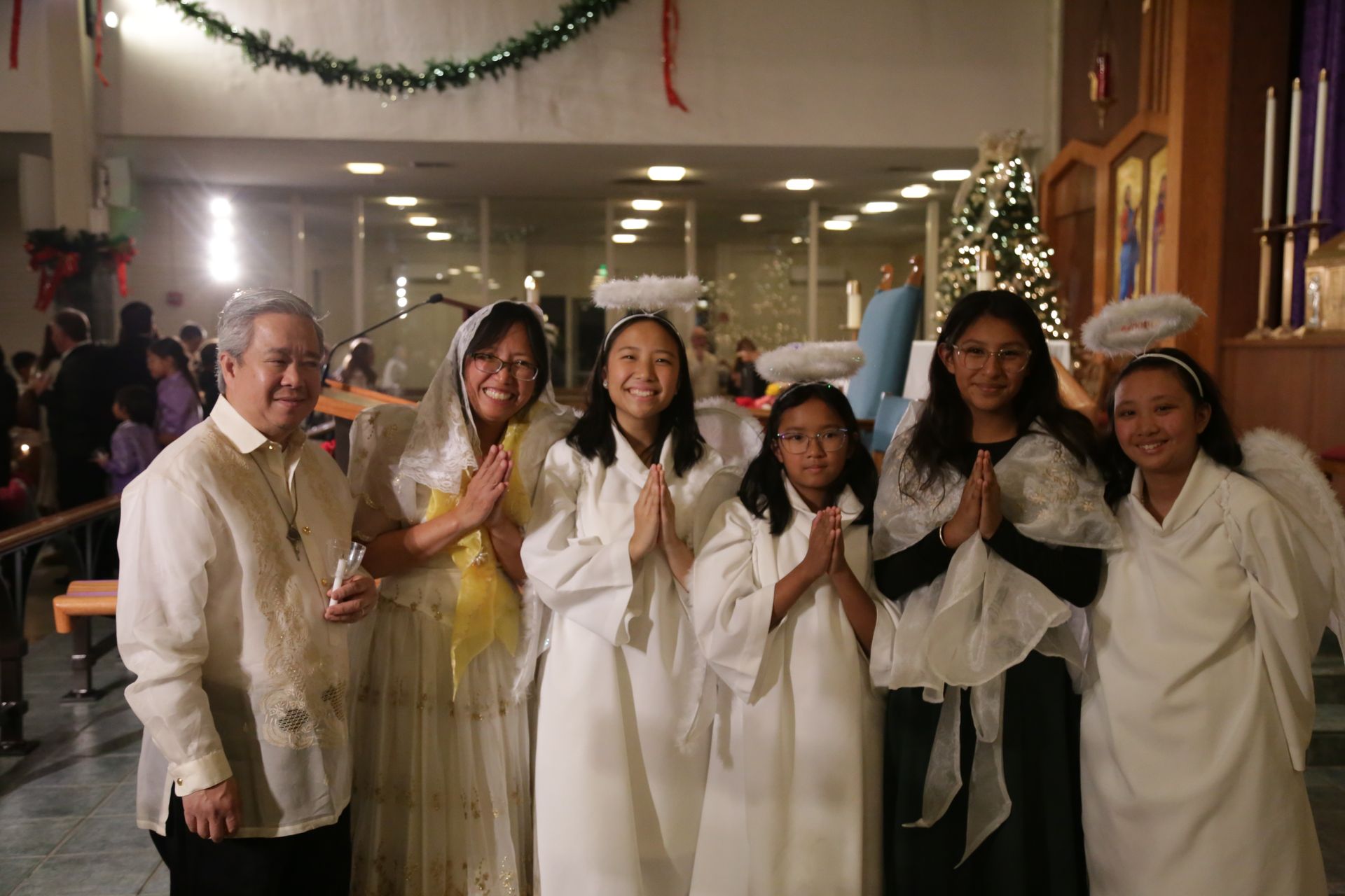 Six people in white attire pose indoors beside a lit Christmas tree.