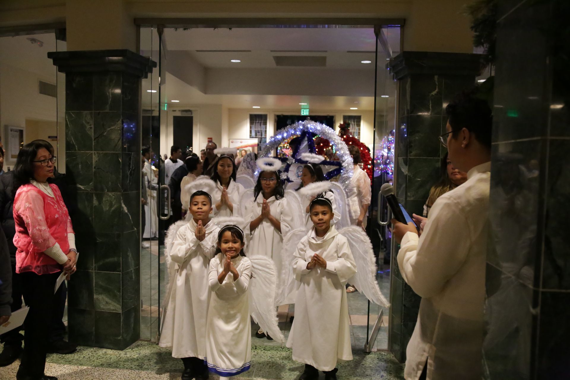 Children in white costumes walk through a decorated entrance during a festive indoor event