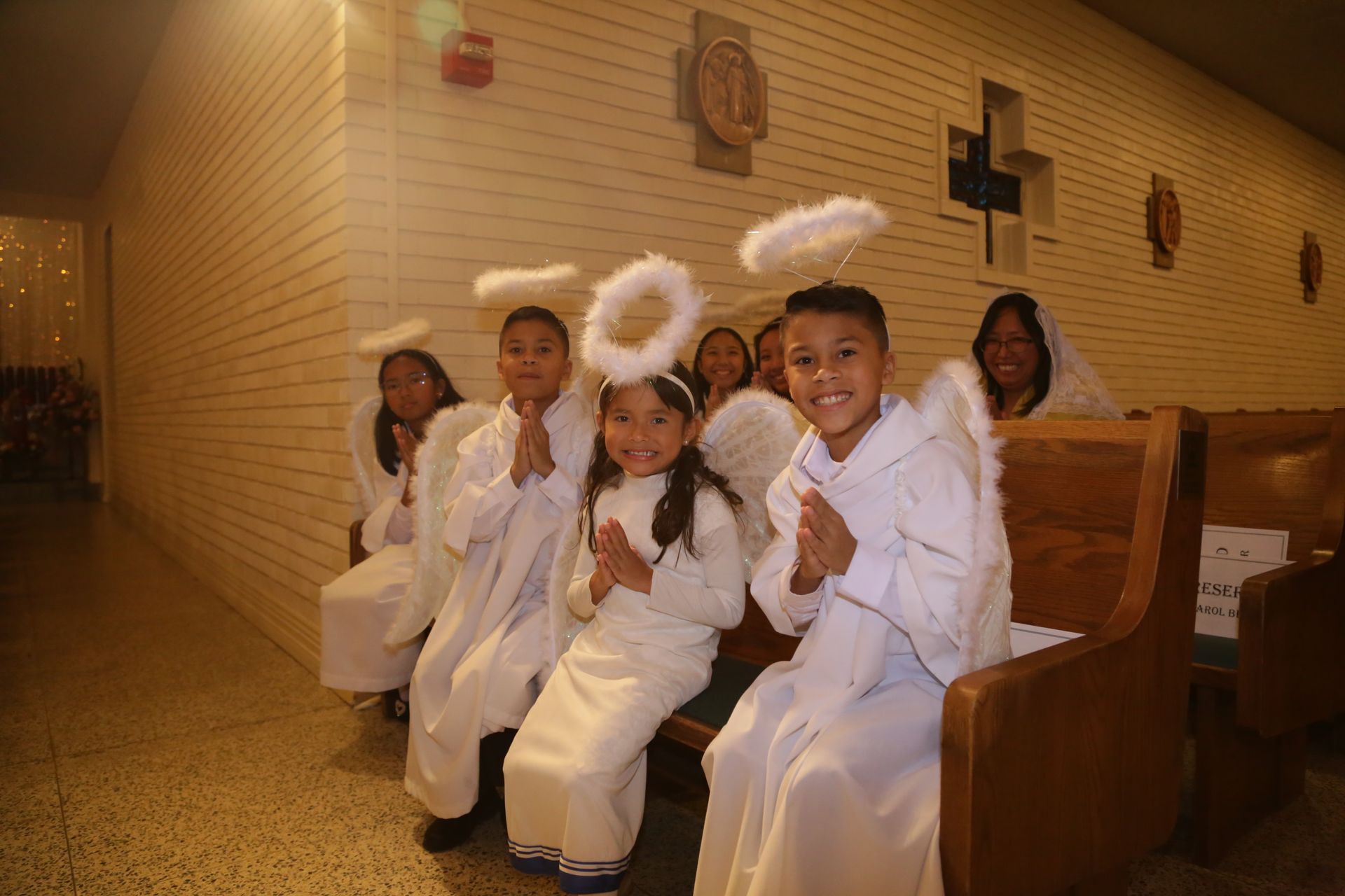 Children in white costumes and angel halos sit on a church pew, smiling in a warm-lit chapel.