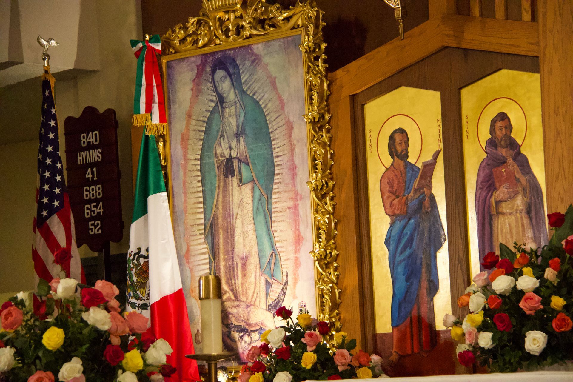Church altar with framed religious icons, floral arrangements, and an Italian flag draped beside them