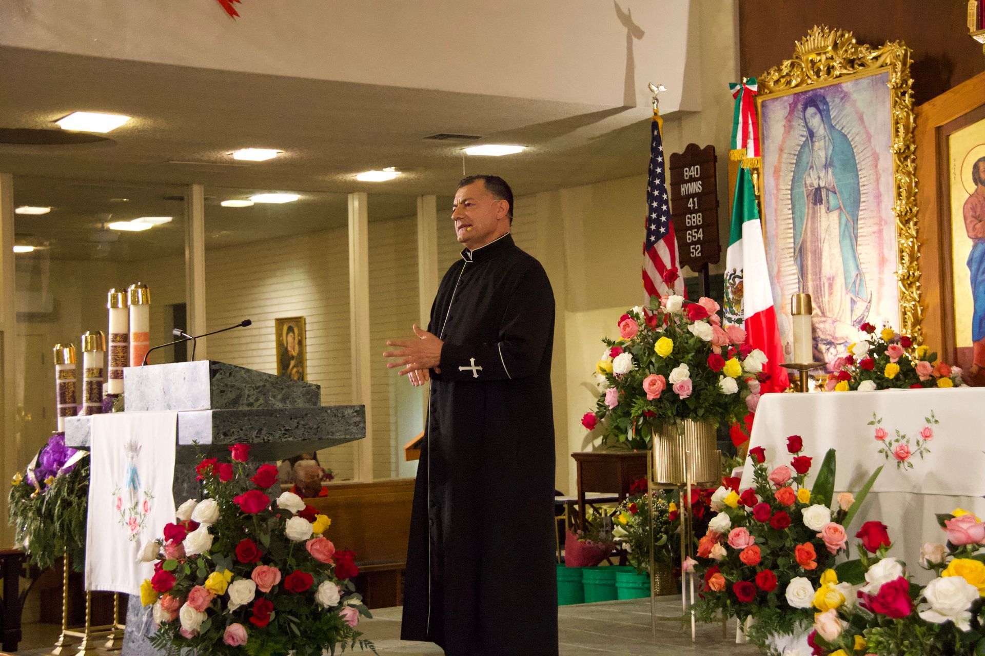 Priest speaking at an altar decorated with flowers beside a religious icon and candlelight