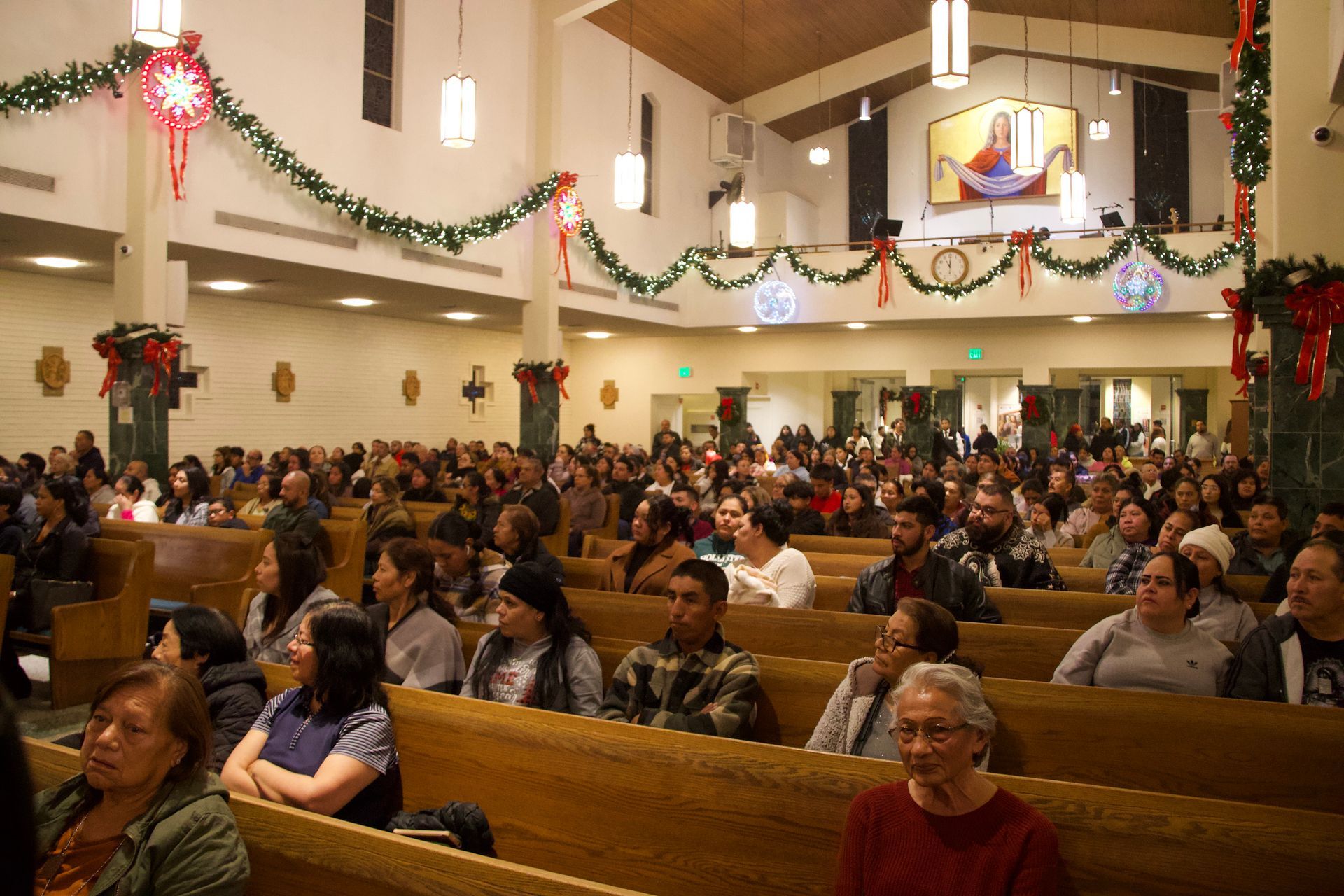 Crowded church hall with people seated on wooden pews during a holiday service, decorated with garlands and wreaths
