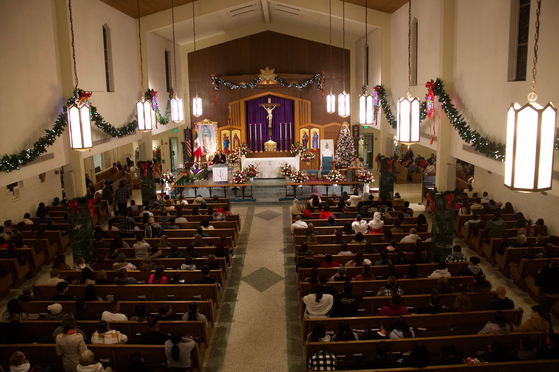 Wide view of a church service with congregants seated in pews facing a lit altar and purple drapery