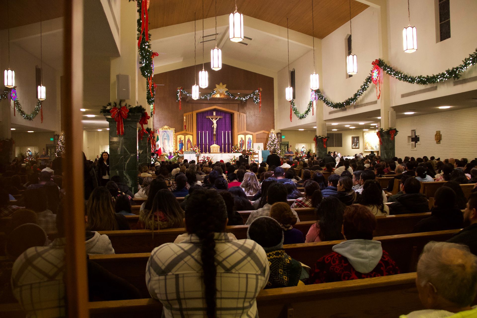 Crowded church service with people seated in pews facing a decorated altar and purple drapery