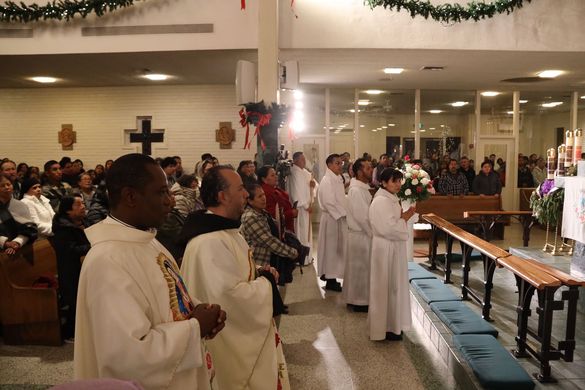People in white robes stand in a church aisle during a religious ceremony, with a crowd seated nearby.