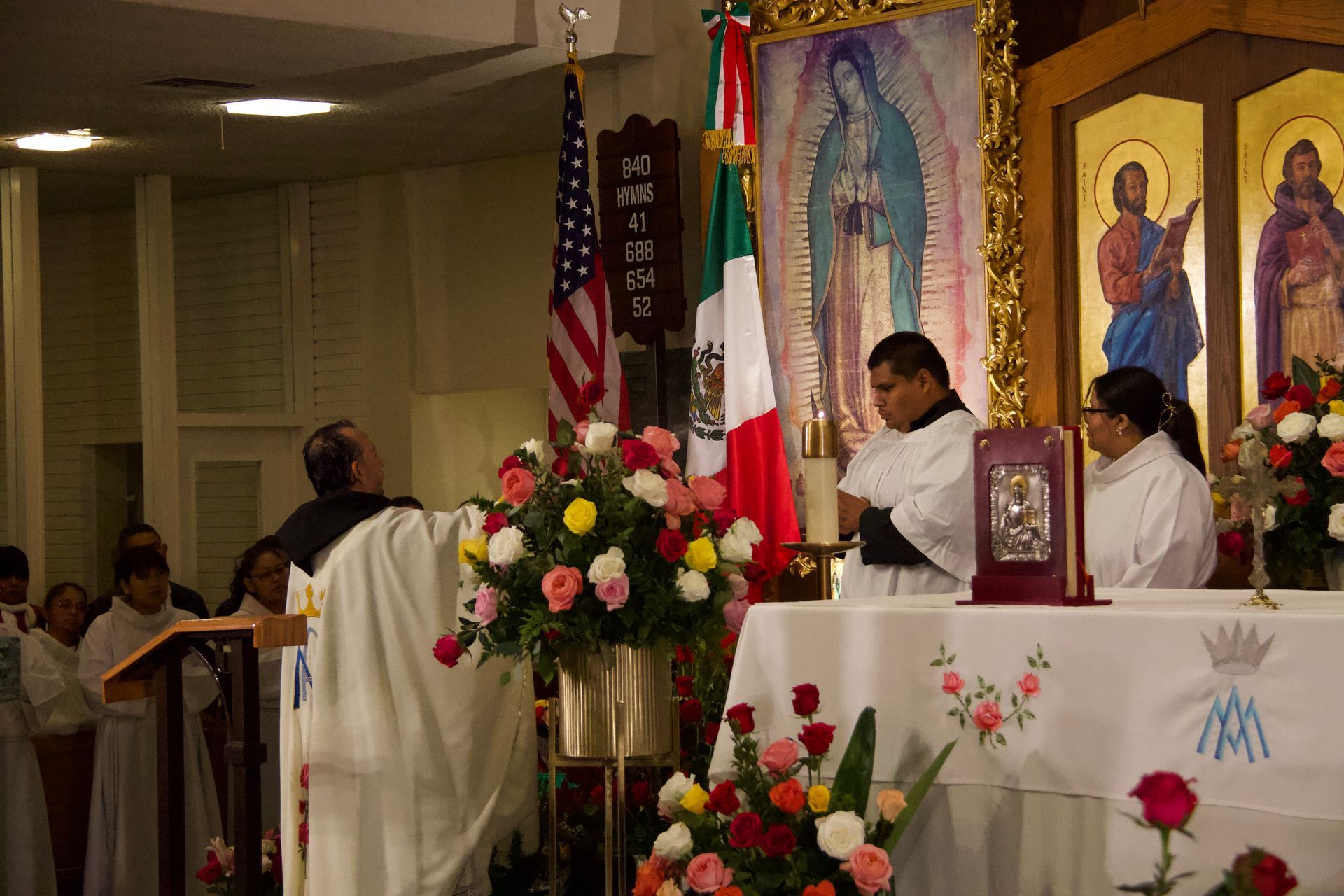 Priest and altar servers at a church altar with flowers, flags, and religious banners during Mass