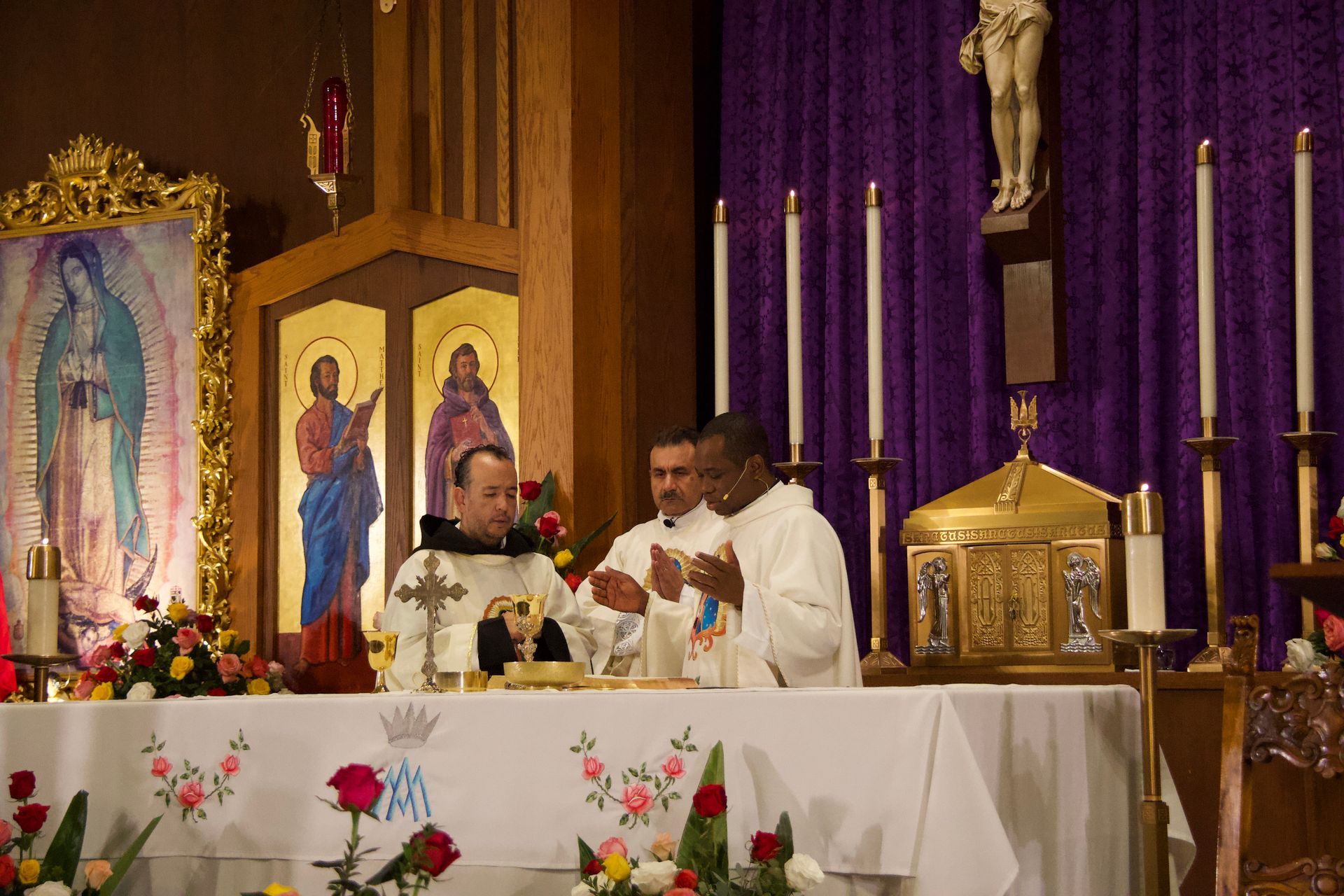 Priests celebrating Mass at a church altar with candles, flowers, and purple drapery