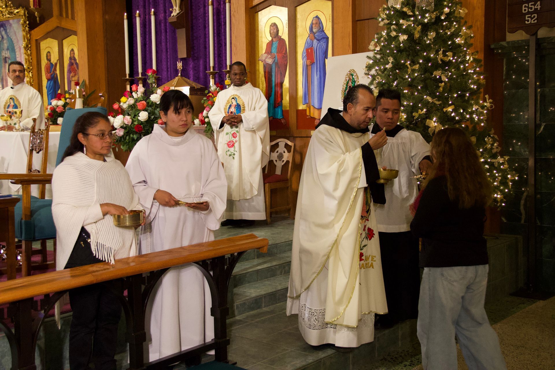 Clergy and altar servers in a church during a Christmas service, with a decorated tree and lit candles.