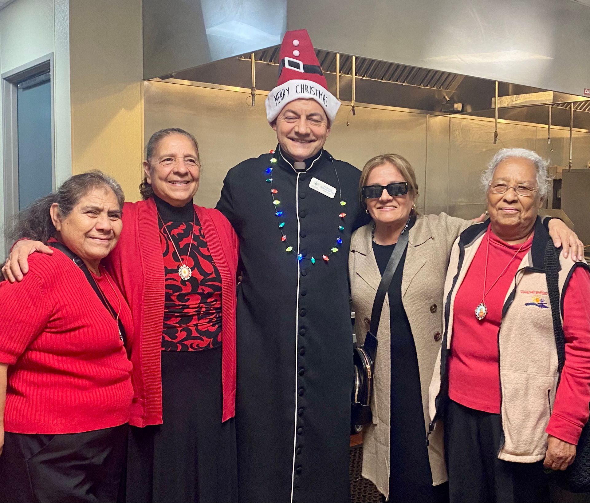 Group of five smiling people posing indoors, one wearing a festive hat and necklace, in front of a kitchen counter.