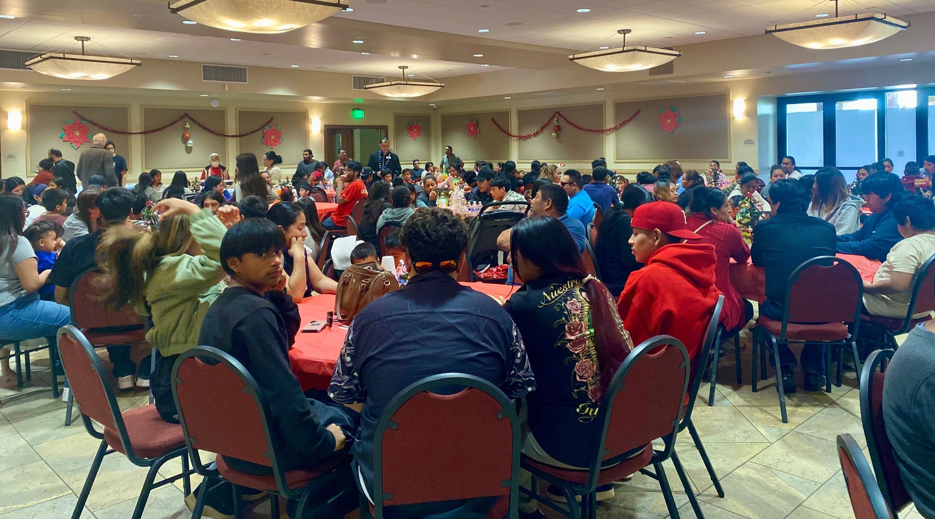 People seated at round tables in a large banquet hall, many wearing red jackets and hats.