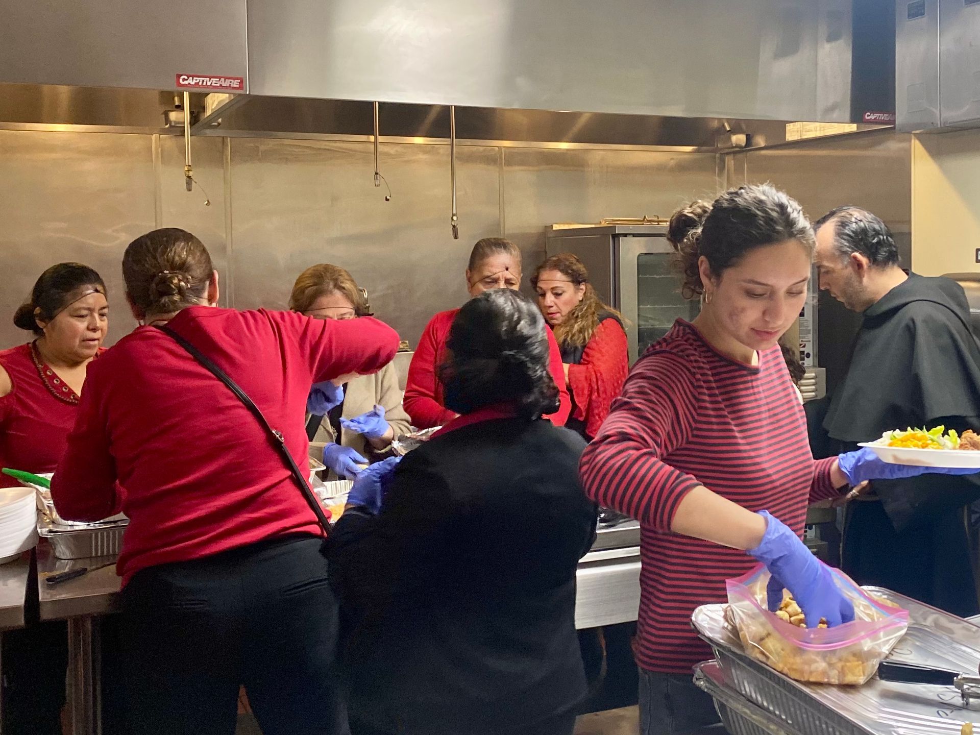 People serving food in a busy kitchen line, with trays of colorful ingredients and stainless-steel counters.