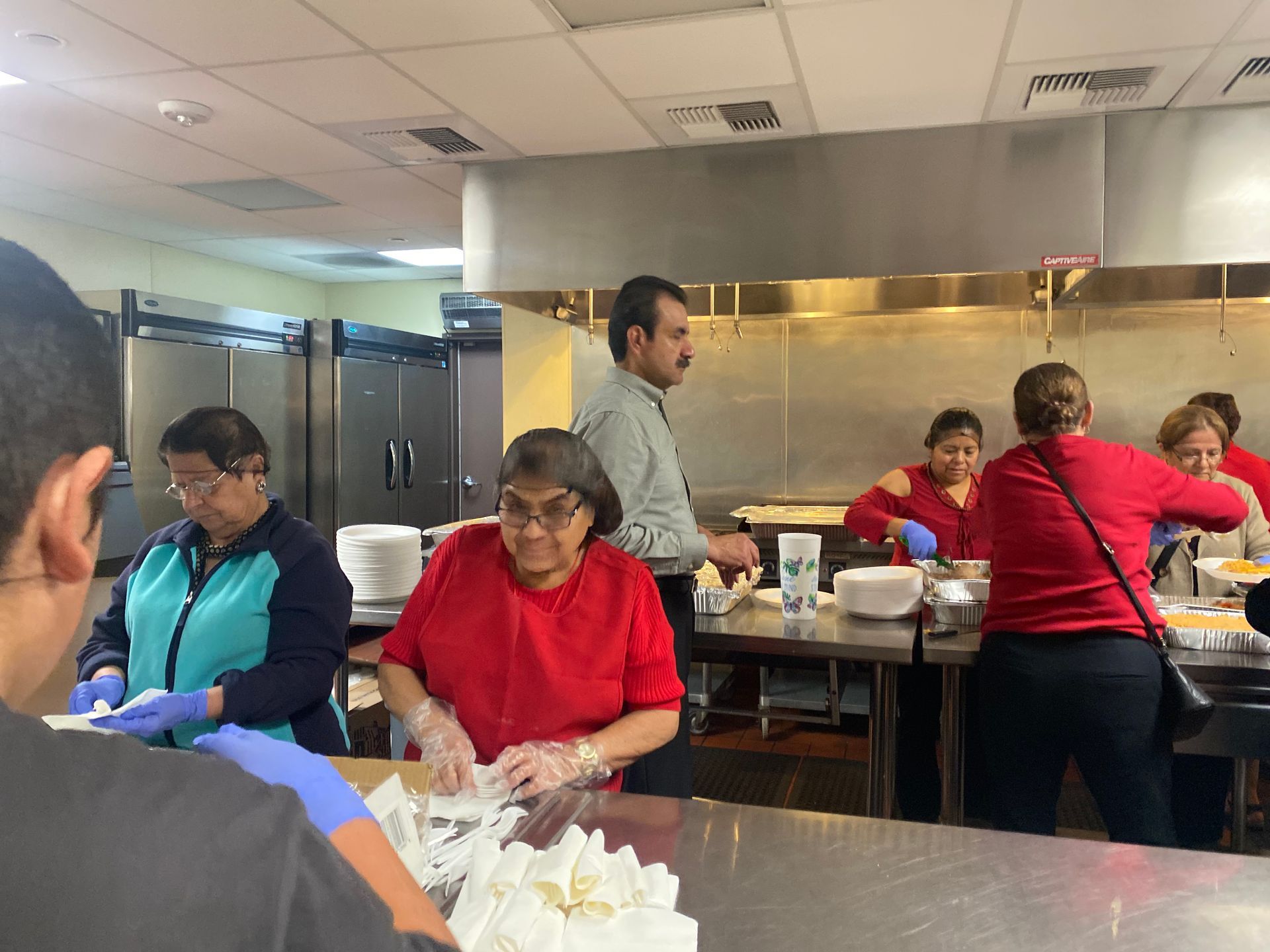 Volunteers preparing and serving food in a kitchen with stainless steel counters and trays.