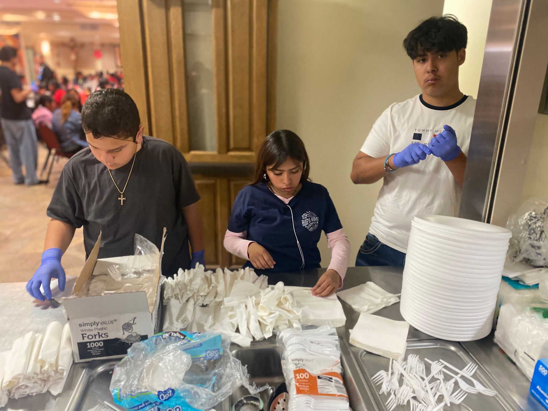 Three volunteers in gloves sorting disposable utensils and plates on a table at an indoor event