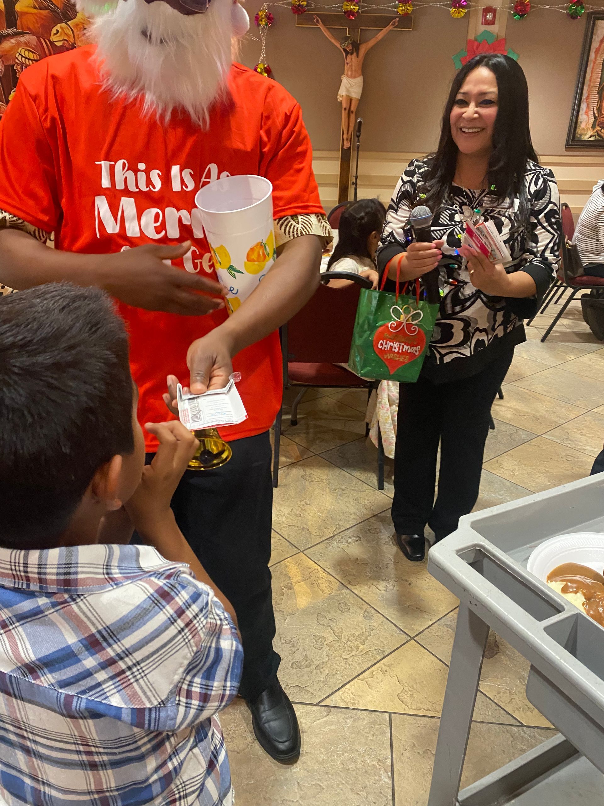 Santa in a red shirt hands out treats to a child beside a woman holding a green gift bag.